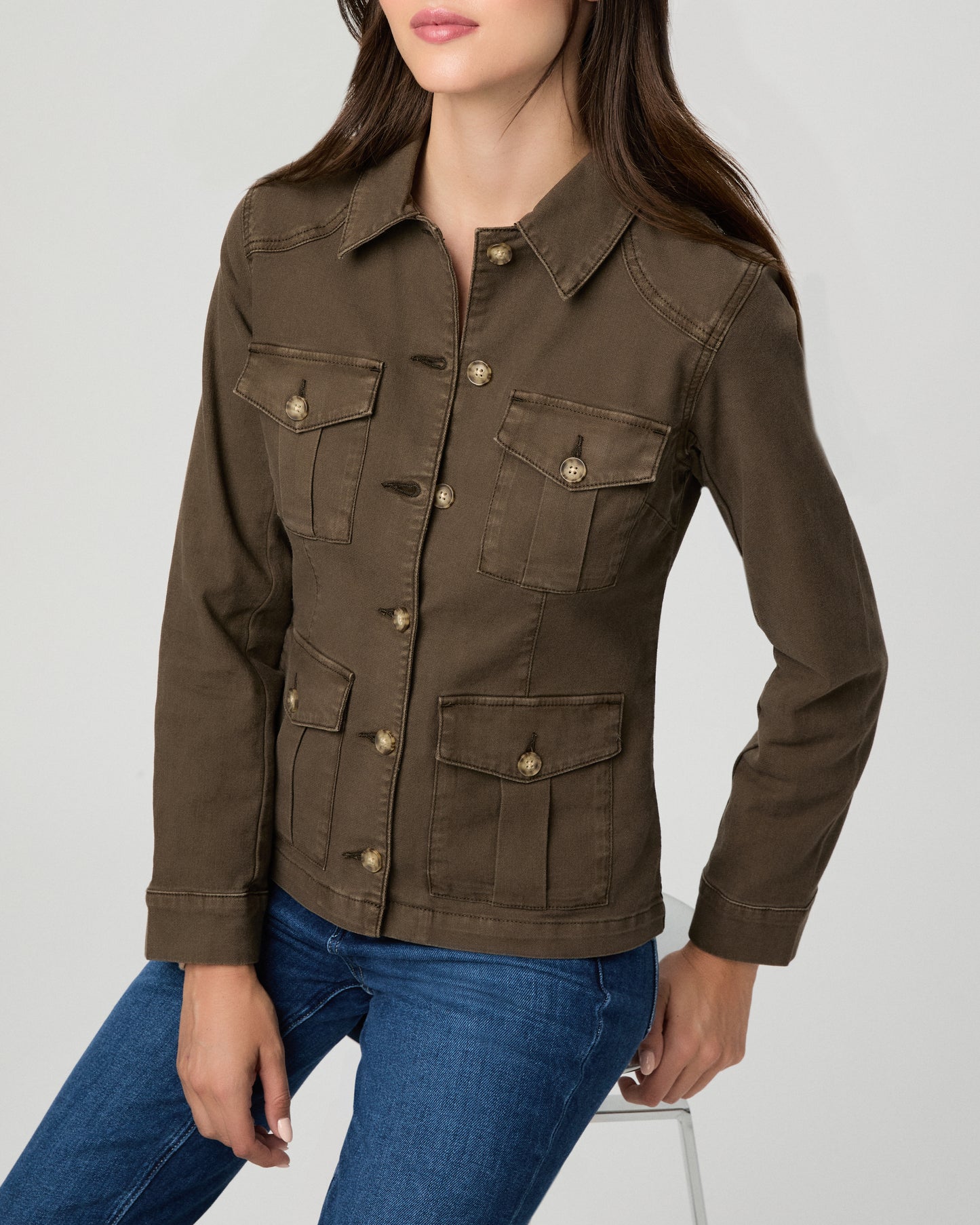 A woman modeling a dark brown utility jacket with four front flap pockets and gold-tone buttons, paired with blue jeans, against a light gray studio background.