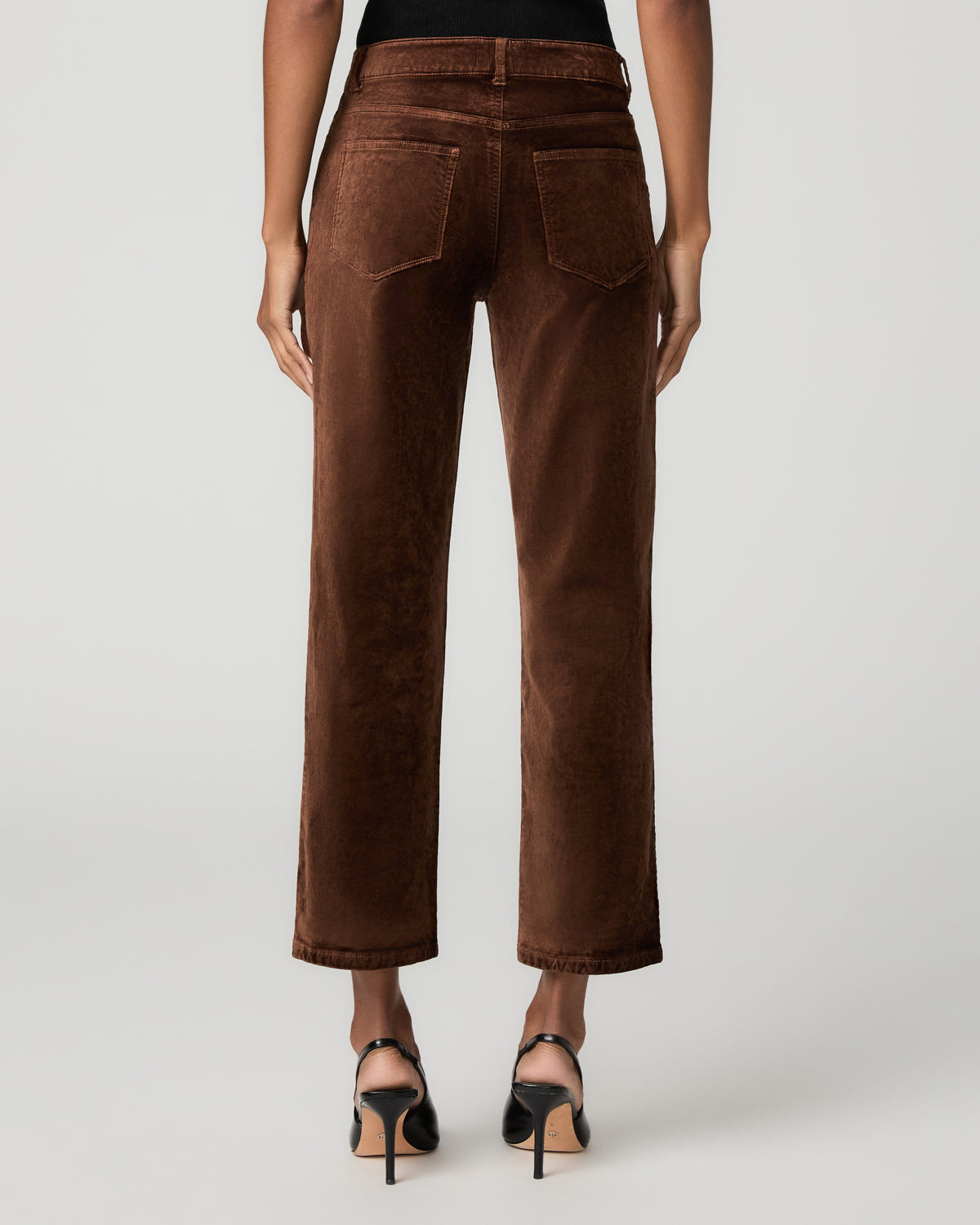 Back view close-up of a woman wearing brown velvet straight-leg ankle jeans with black ankle-strap heels.