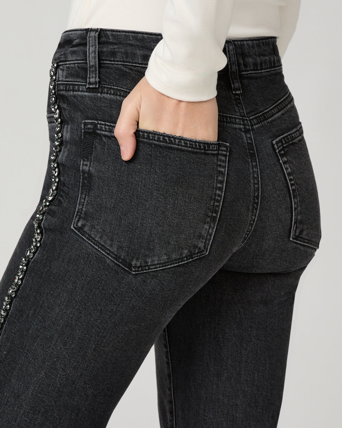 Close-up of the washed black Mason Ankle Straight Jean's back pocket and embellished side seam, with a hand tucked into the pocket.