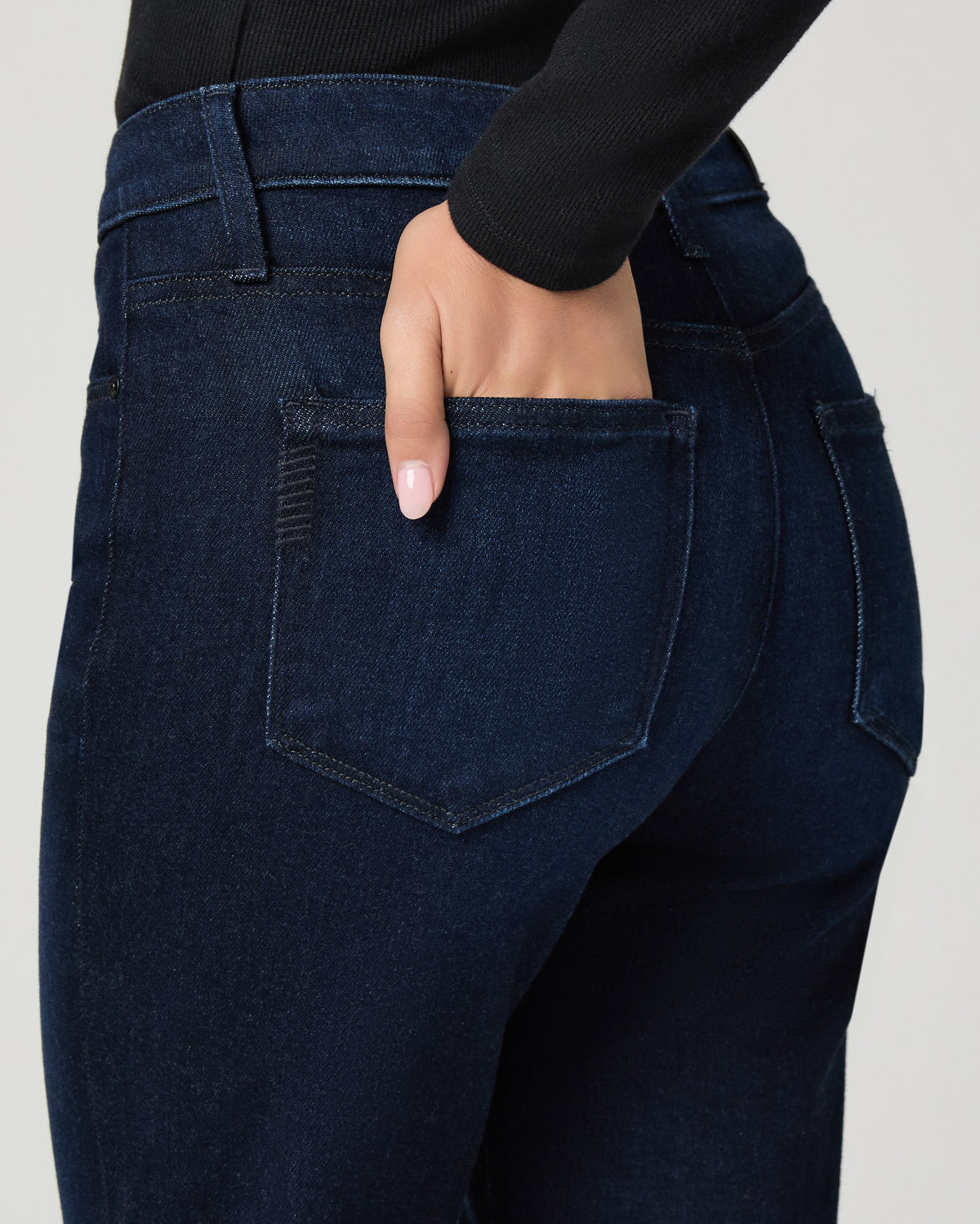 Close-up of a person wearing dark indigo denim jeans, showing the back pocket and hand in the pocket, paired with a black long-sleeve top against a light gray background.