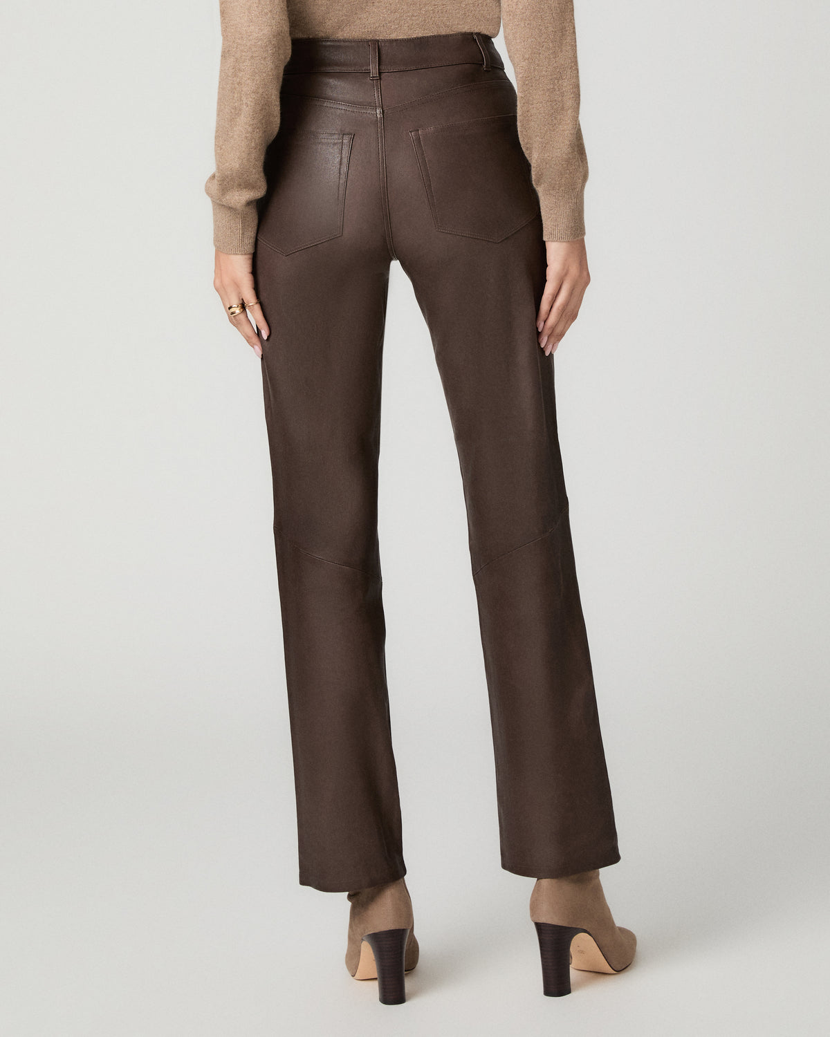 A model shown from the back wearing dark brown high-waisted straight-leg trousers, a beige knit sweater, and beige high-heeled shoes against a light gray studio background.