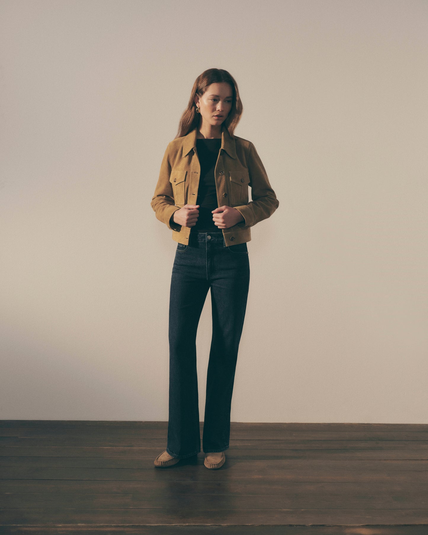 Person wearing a brown jacket and dark jeans standing against a plain background