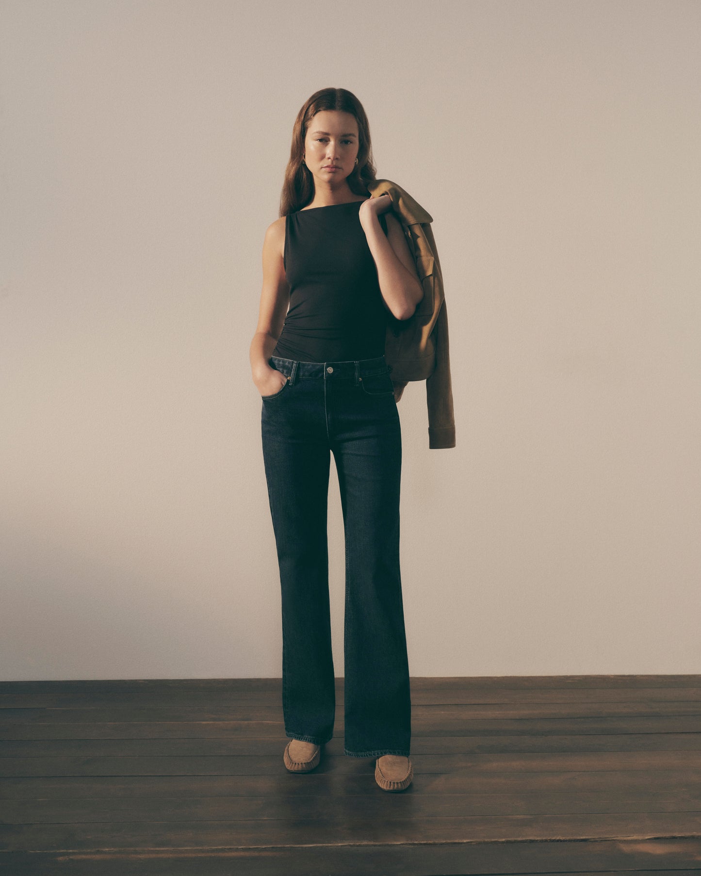 Woman wearing a black sleeveless top and dark jeans, holding a brown jacket against a plain background.