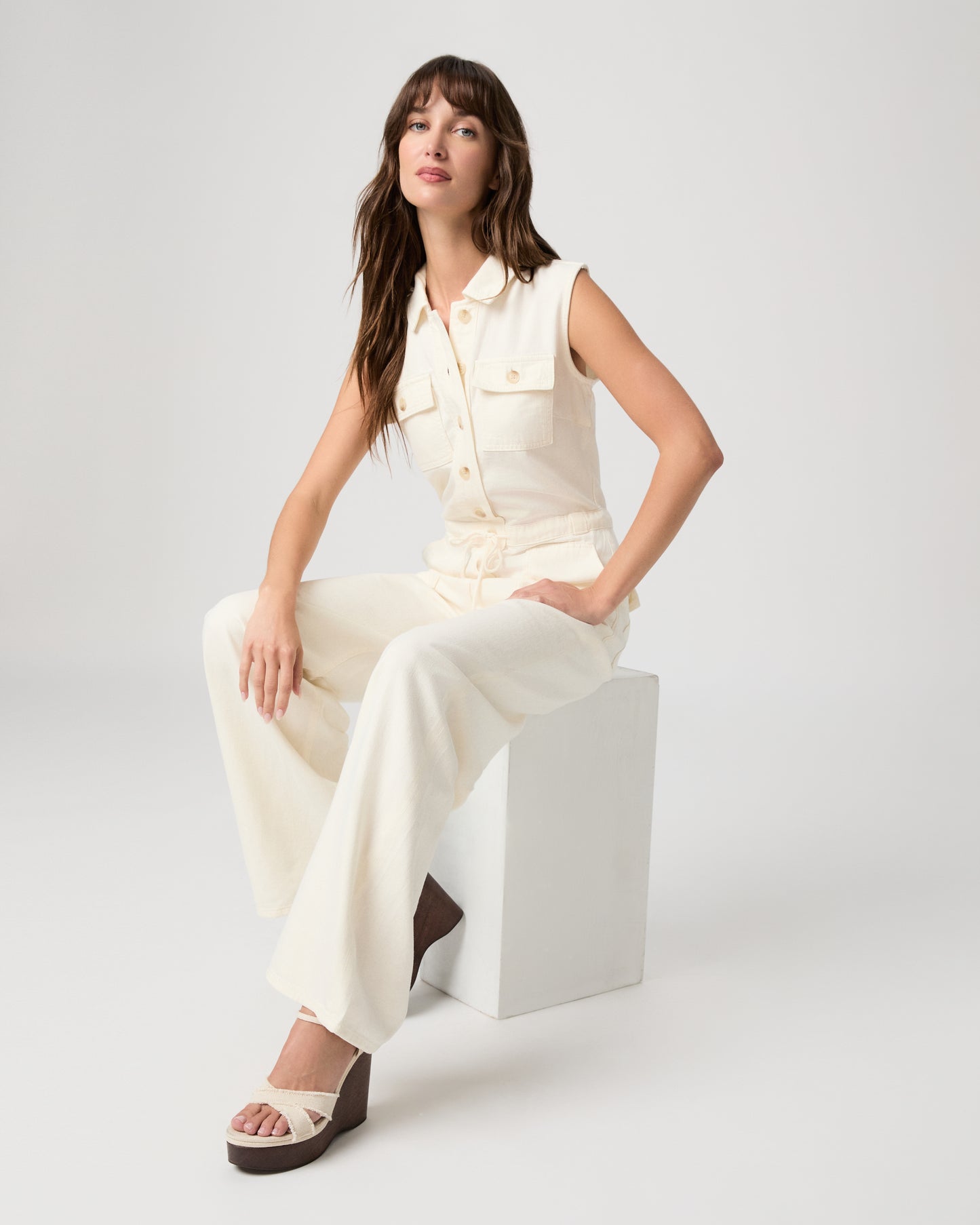 Woman sitting on a white stool wearing a white sleeveless jumpsuit on a plain background