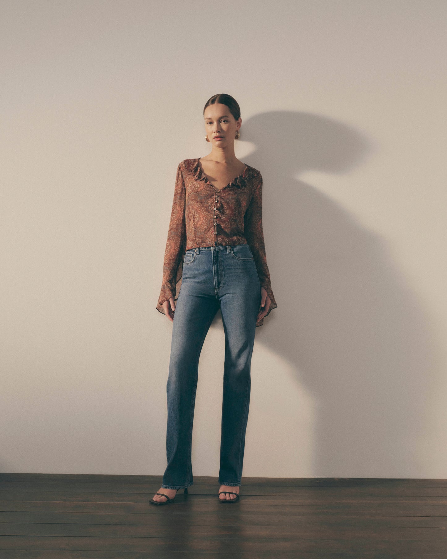 Woman wearing a patterned blouse and blue jeans standing against a plain wall.