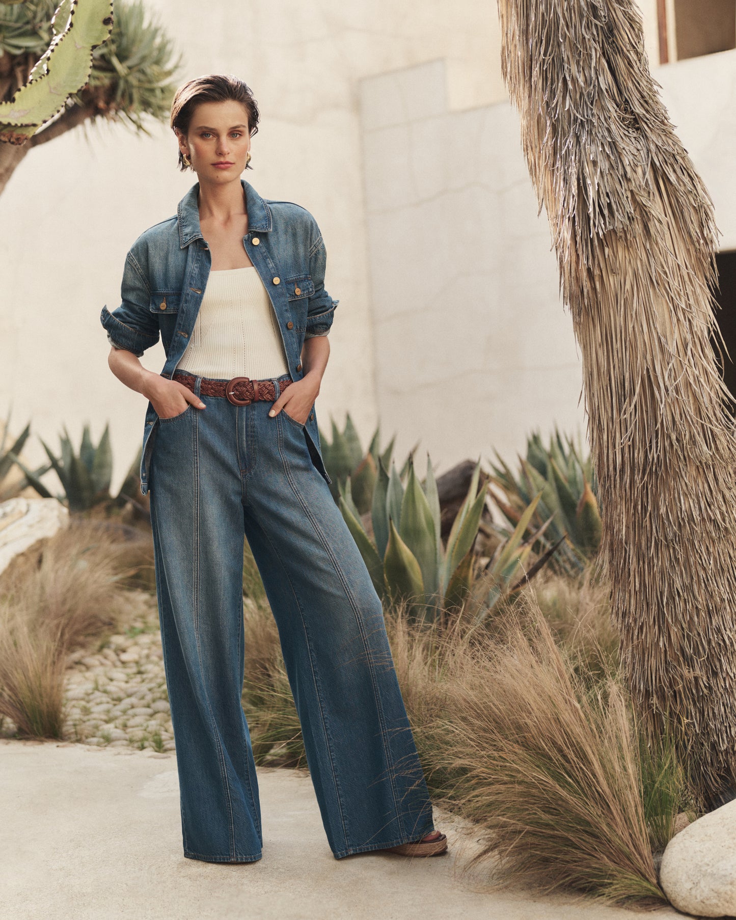 Woman wearing a denim outfit standing in a desert-like setting with cacti and dry grass.