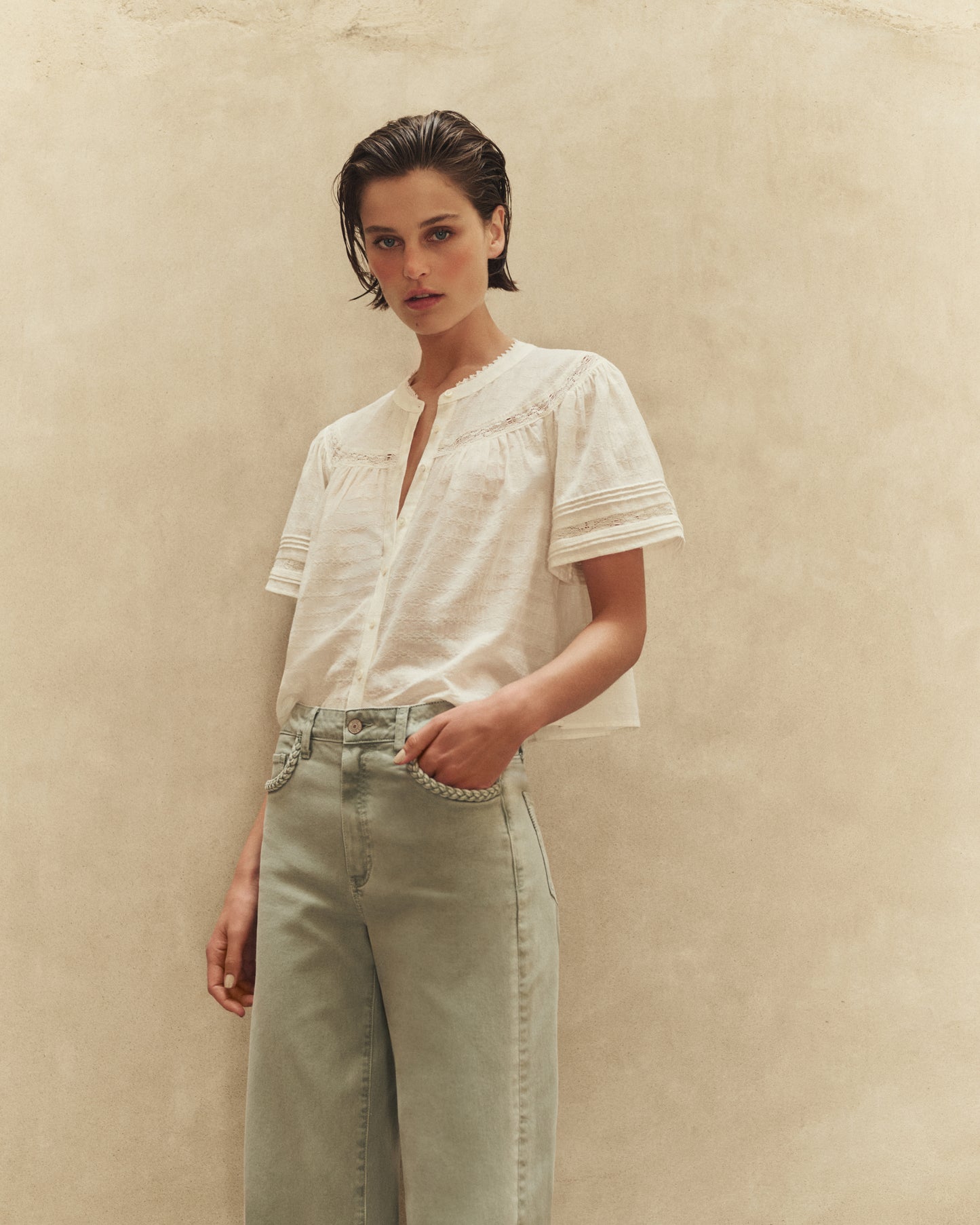 Woman wearing a white blouse and light green pants against a beige background