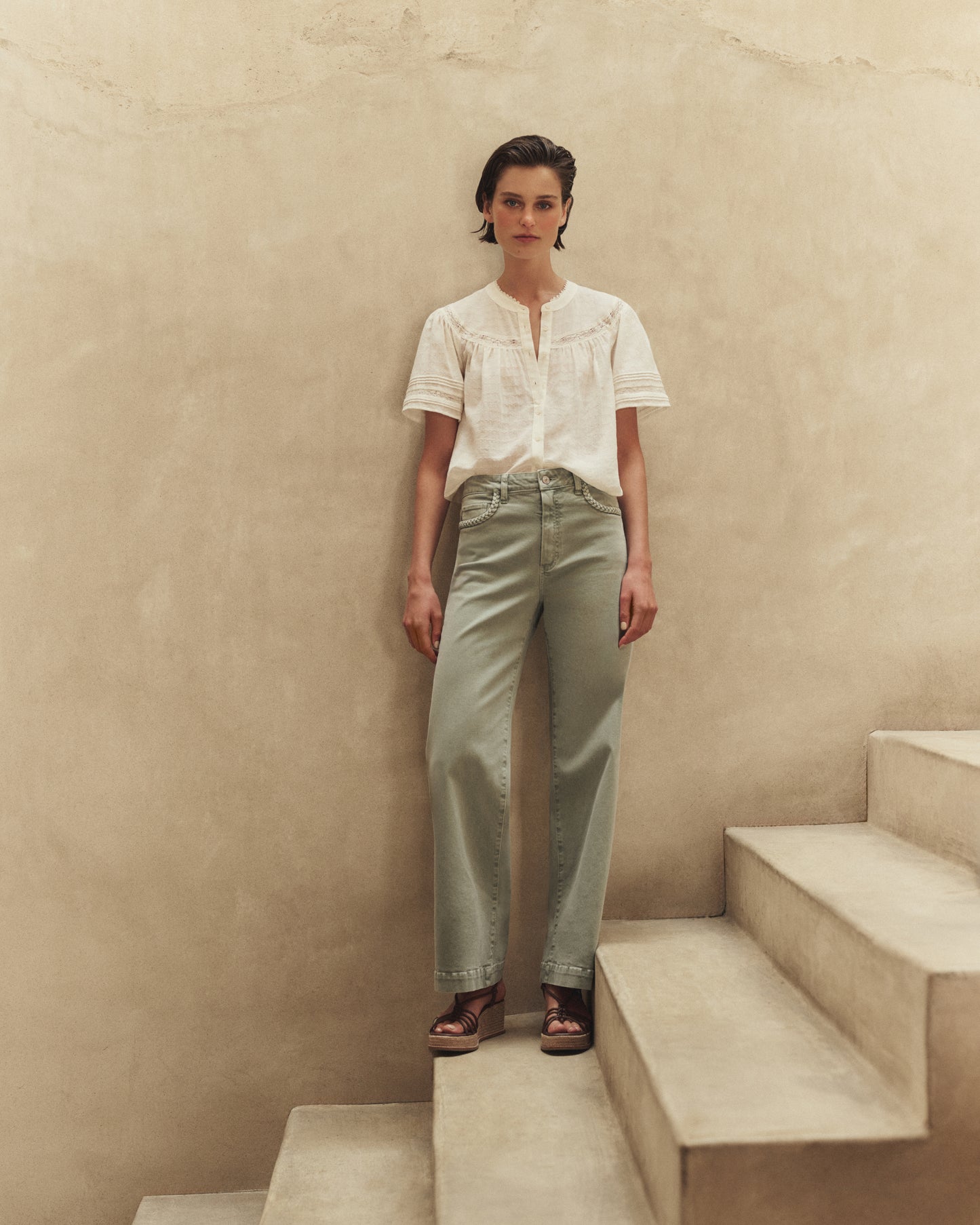 Woman standing on a set of stairs wearing a white blouse and green pants against a beige wall.