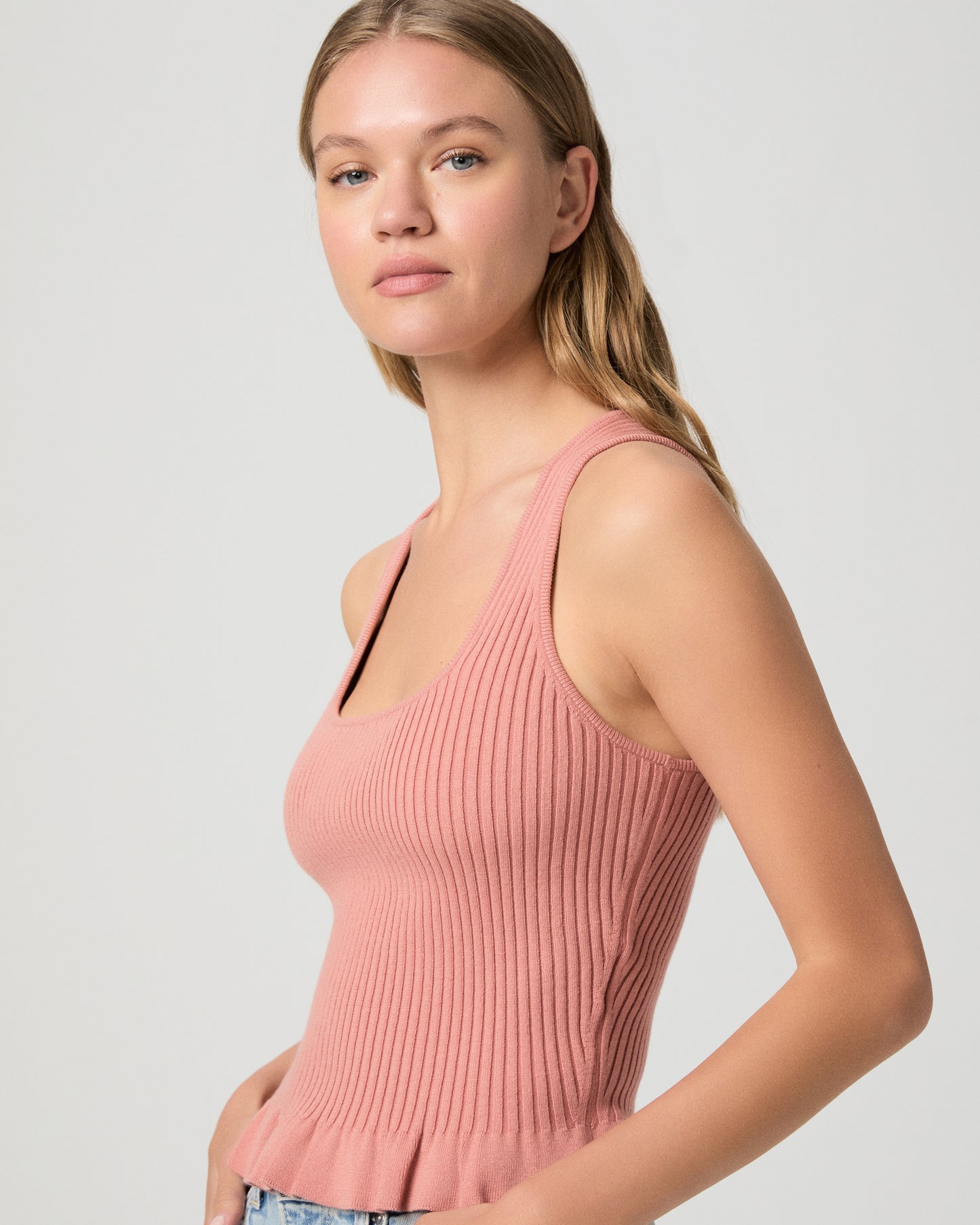 Woman wearing a pink ribbed tank top and light blue jeans on a plain background