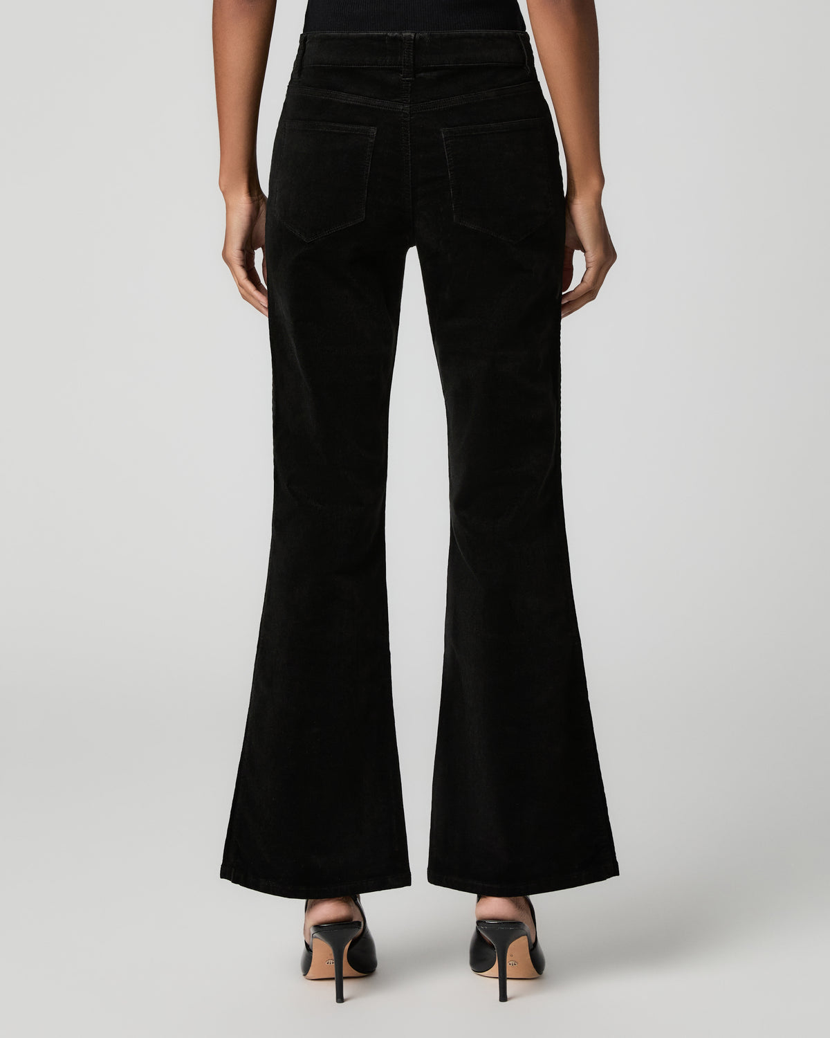 Close-up back view of black corduroy flare pants showing a back patch pocket, paired with black ankle-strap heels.
