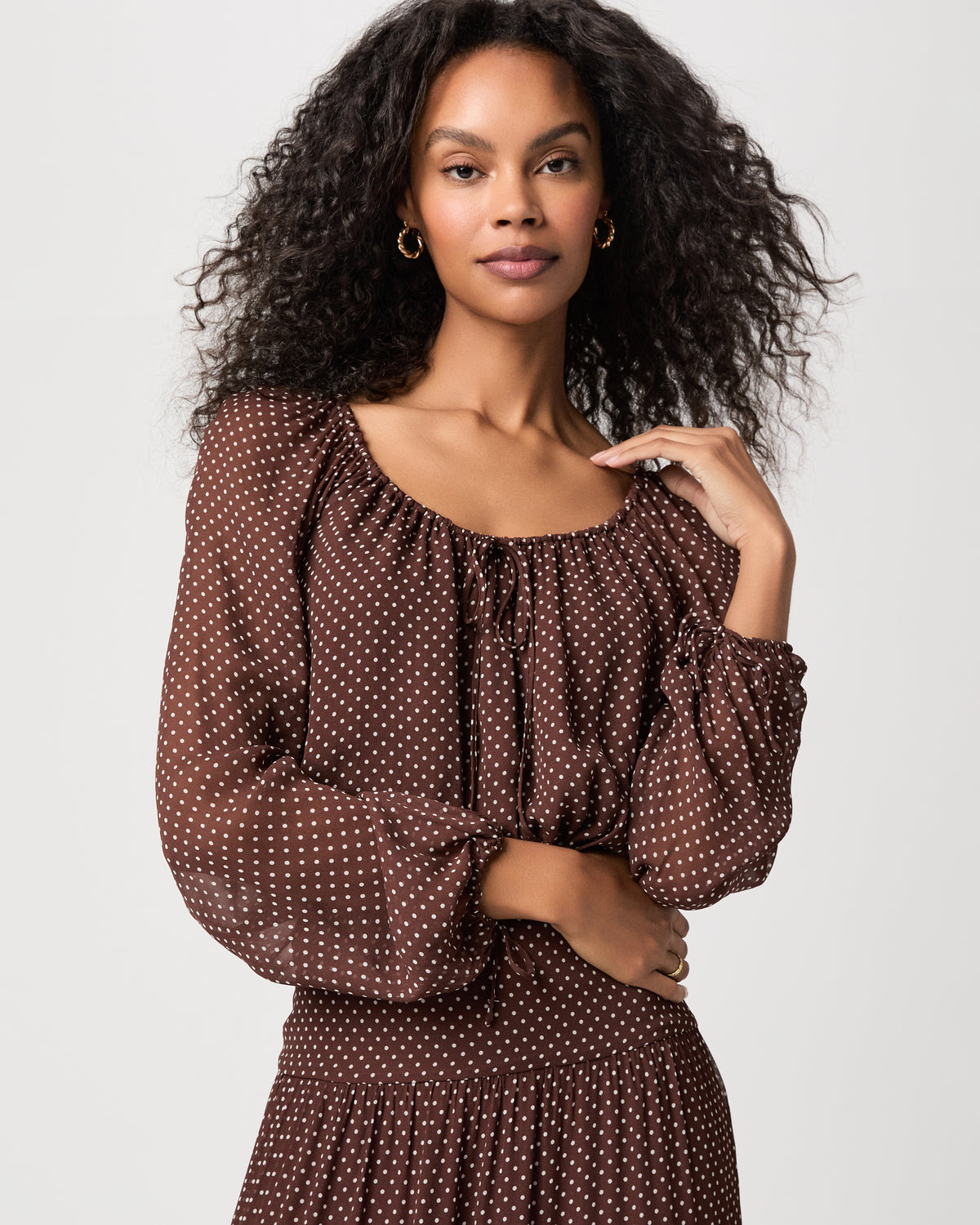 A woman modeling a brown long-sleeve brown polka dot blouse with a matching midi skirt and brown sandals against a light white background.
