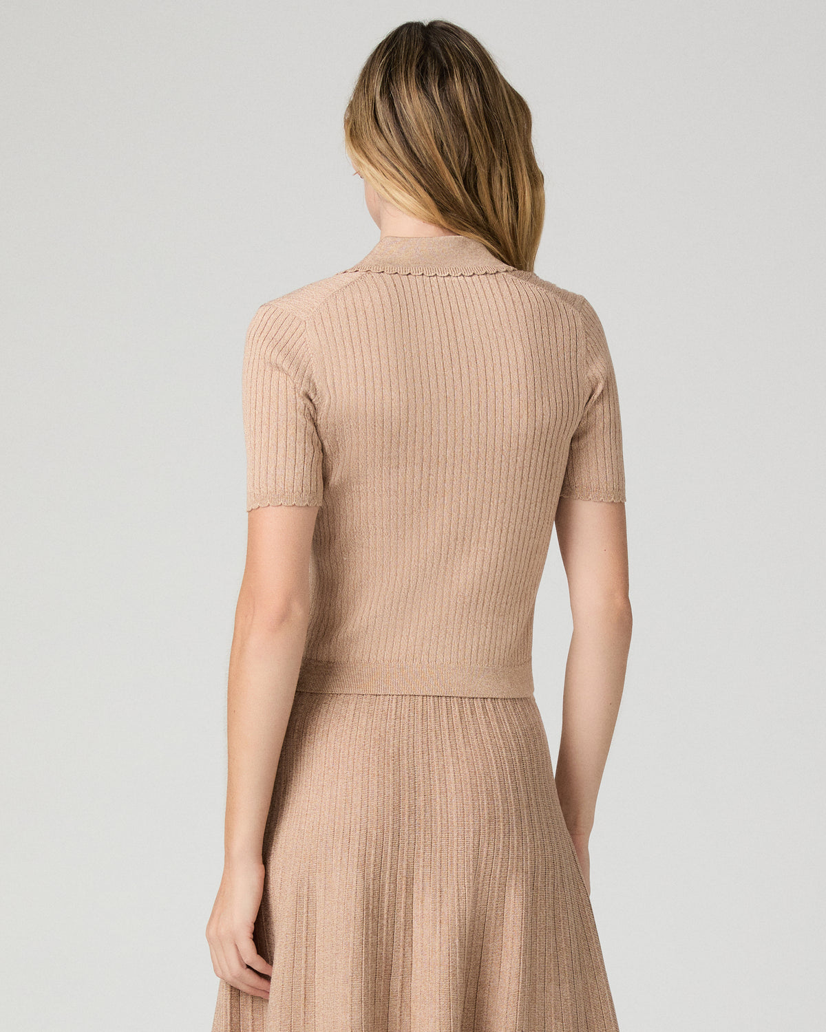 A woman viewed from the back wearing a beige ribbed short-sleeve knit top and matching pleated skirt against a light gray studio background.