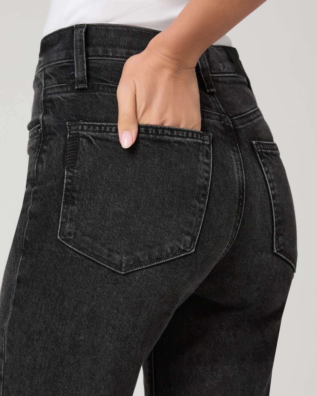 Close-up of a person wearing dark gray washed denim jeans, showing the back pocket and hand in the pocket, against a light gray studio background.