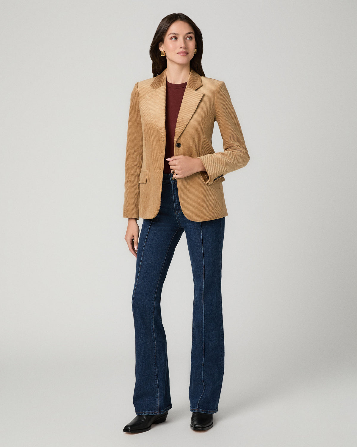 A woman modeling a tan suede-like blazer over a burgundy top and blue flare jeans, standing against a light gray studio background.