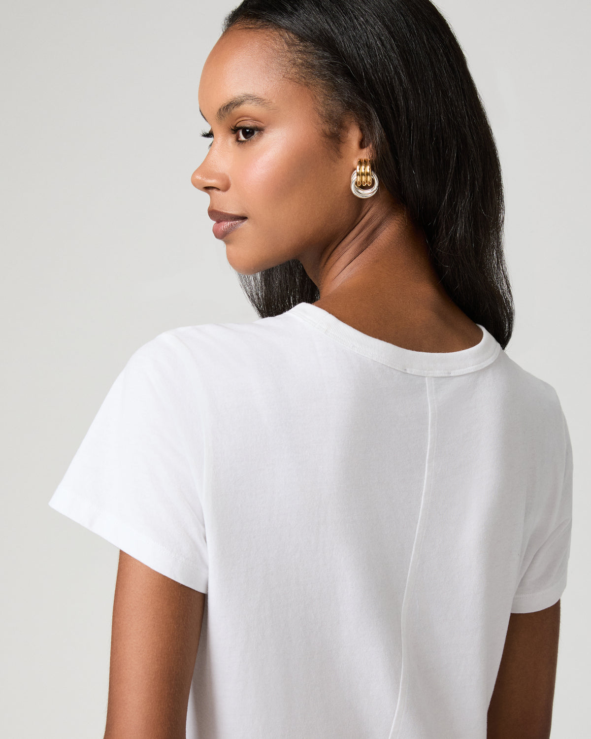 Close-up profile view of a woman in a white tee, gold hoop earrings visible, looking over her shoulder.