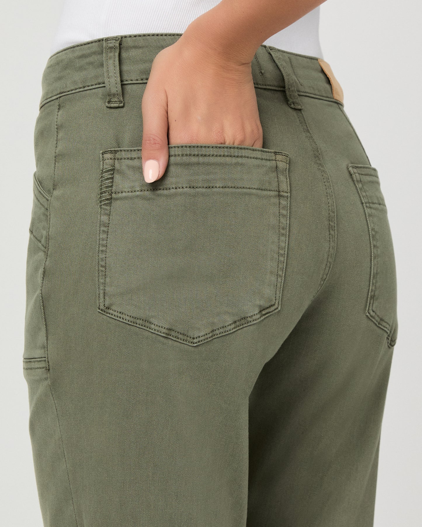 A woman modeling a pair of olive green cropped pants with a white tank top and brown sandals, standing against a light gray studio background.