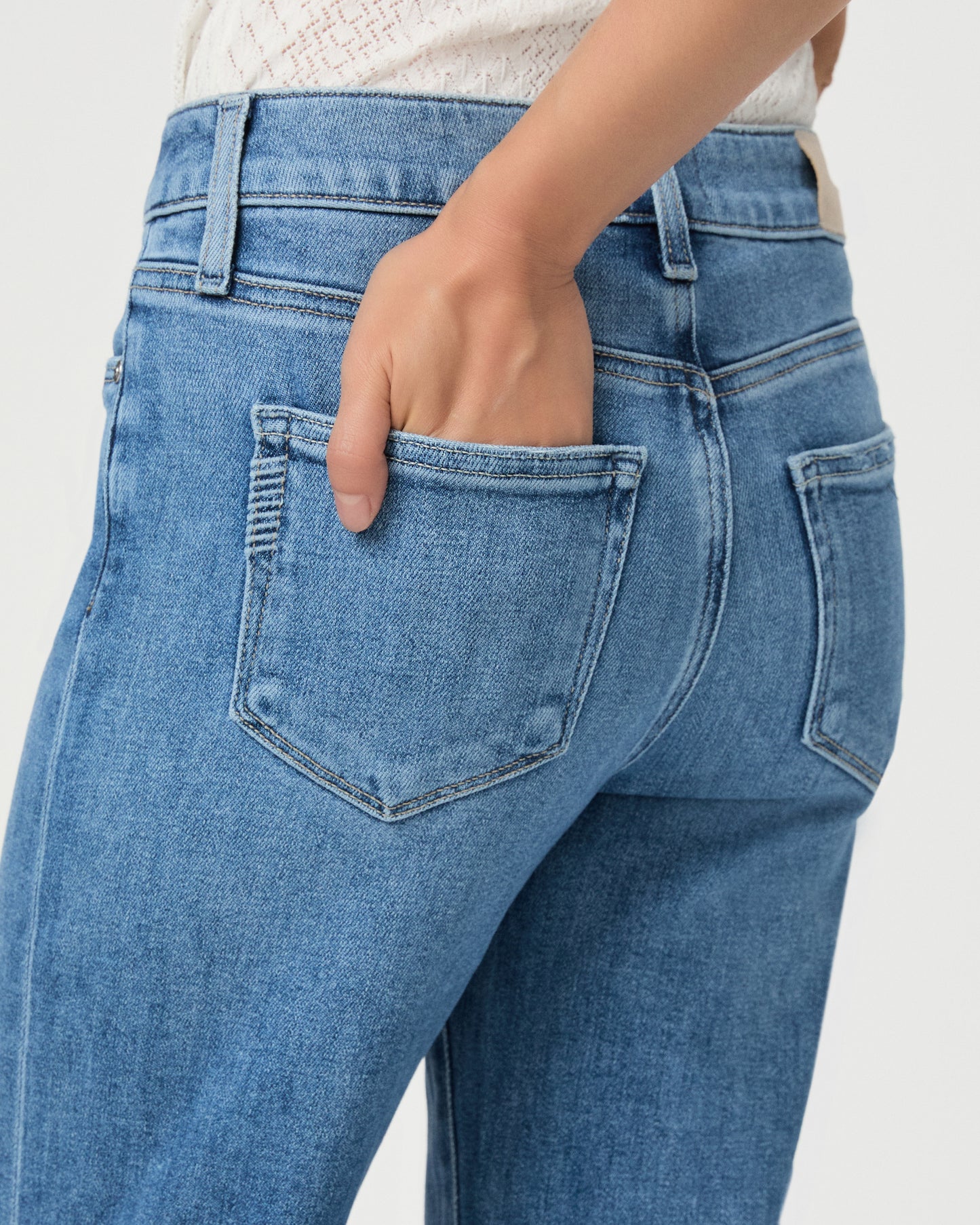 A woman modeling a white textured sleeveless top with ruffle details and high-waisted, wide-leg blue jeans against a light gray studio background.