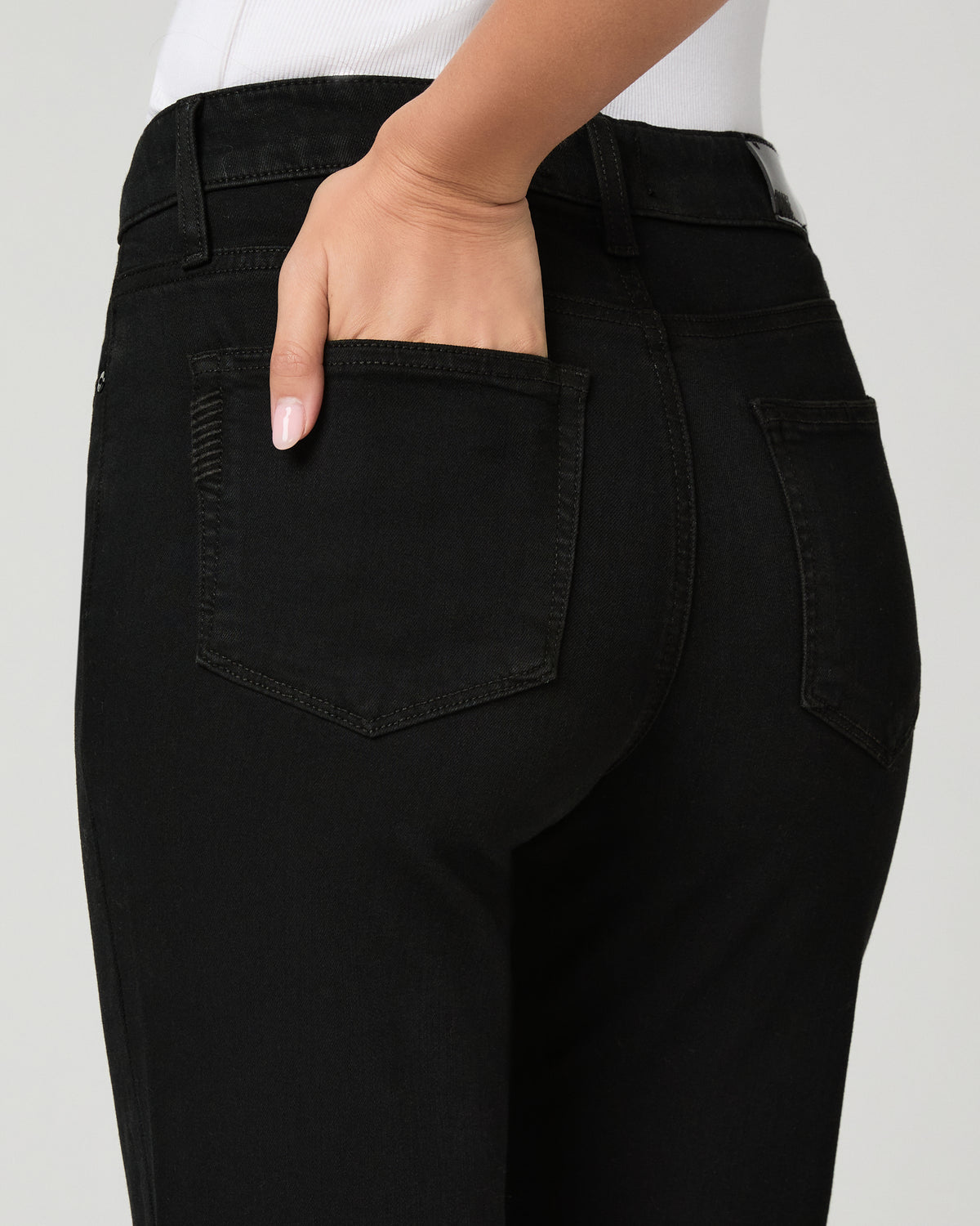 Close-up back view of a person wearing black jeans, with one hand tucked into the back pocket, against a light gray studio background.