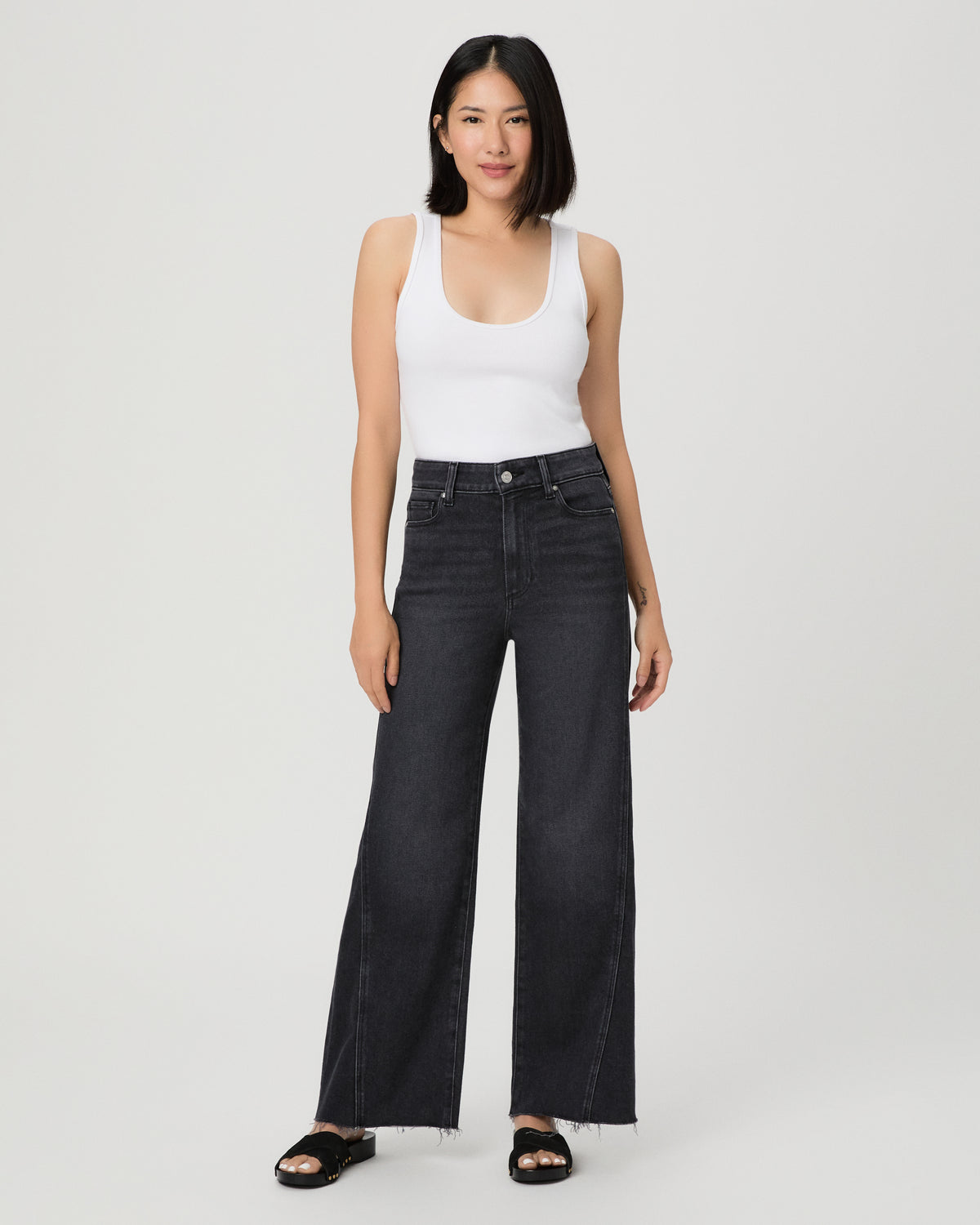 A woman modeling a pair of dark charcoal wide-leg jeans with a frayed hem, paired with a white tank top and black slide sandals, against a light gray studio background.