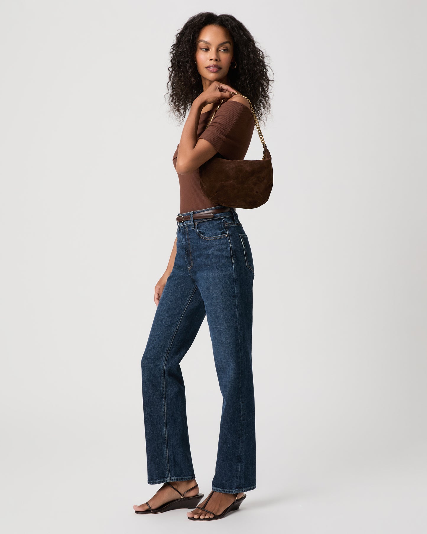 Women wearing brown top paired with blue jeans holding a brown green bag against a white background