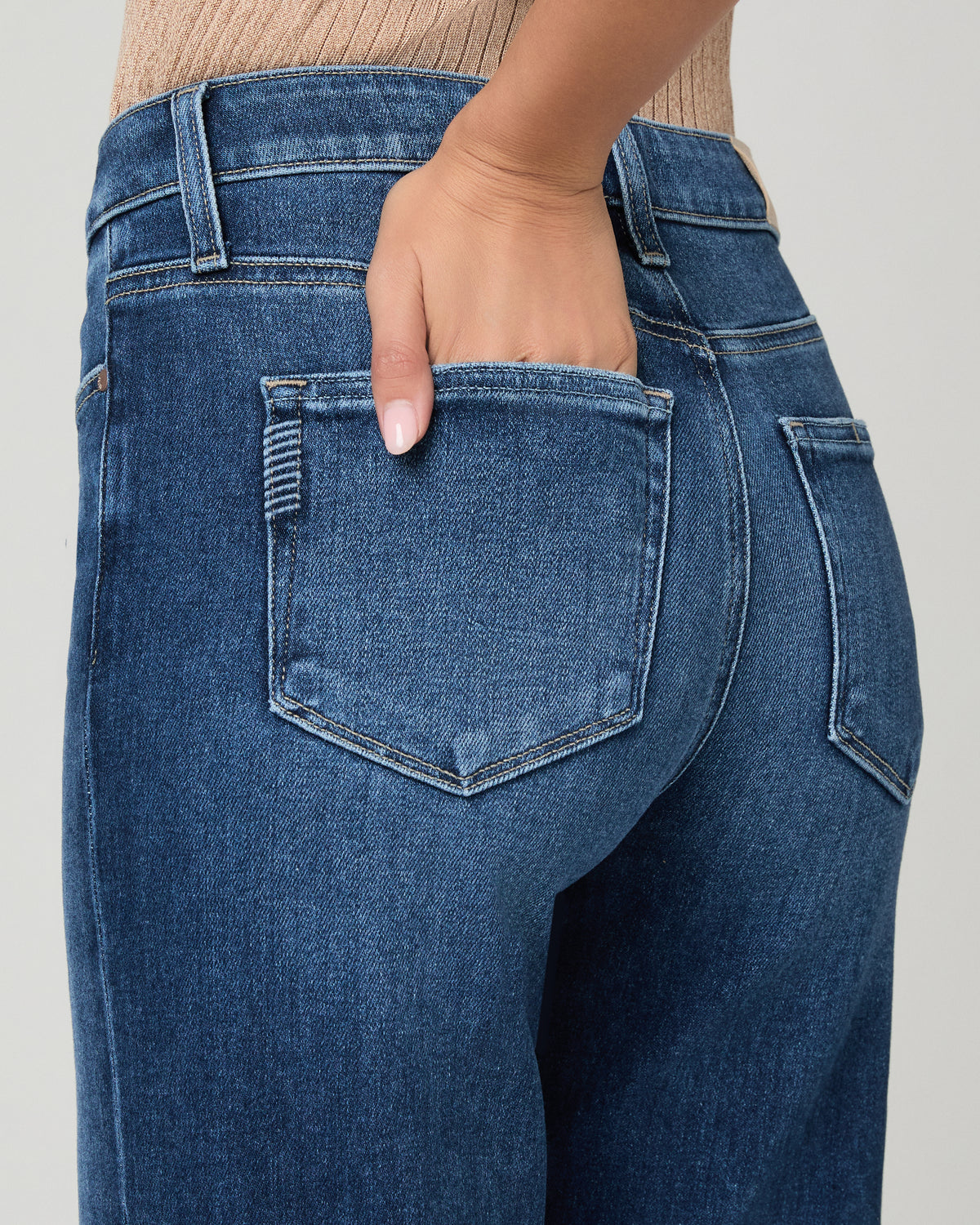 Close-up back view of a person wearing blue denim jeans, showing the back pocket, waistband, and hand in the pocket against a light gray studio background.