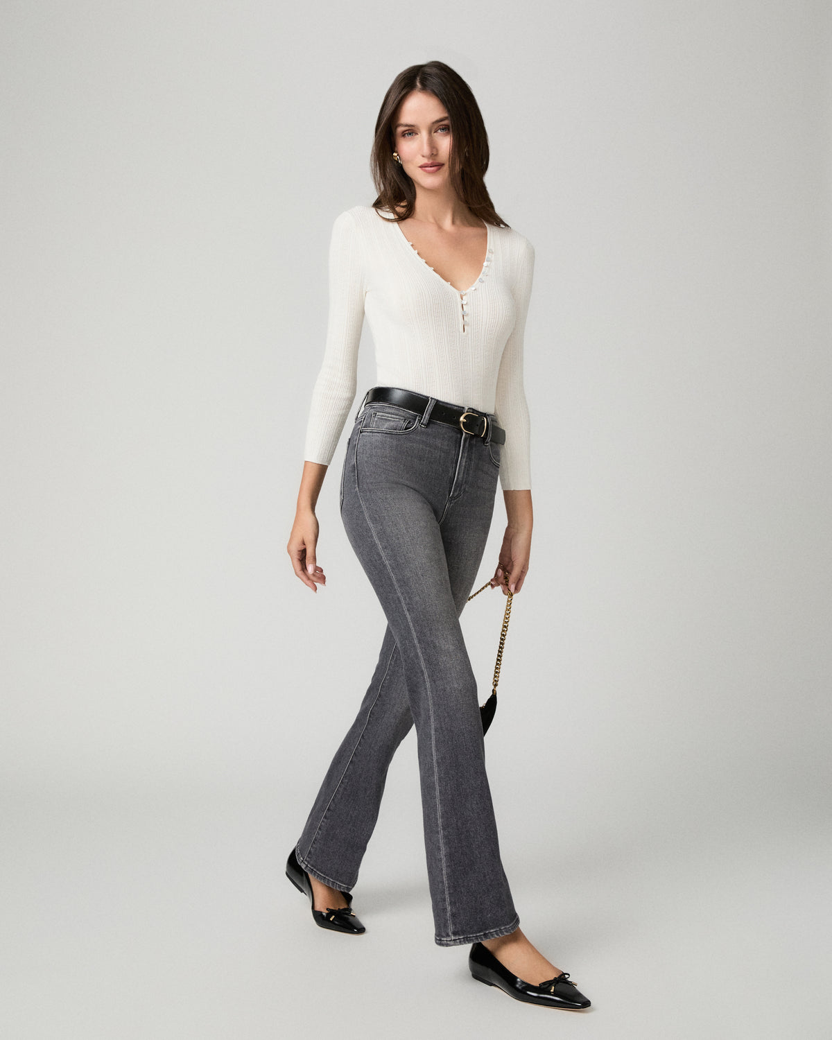 A woman modeling a white long-sleeve knit top and dark gray flared jeans with a black belt, standing against a light gray studio background.