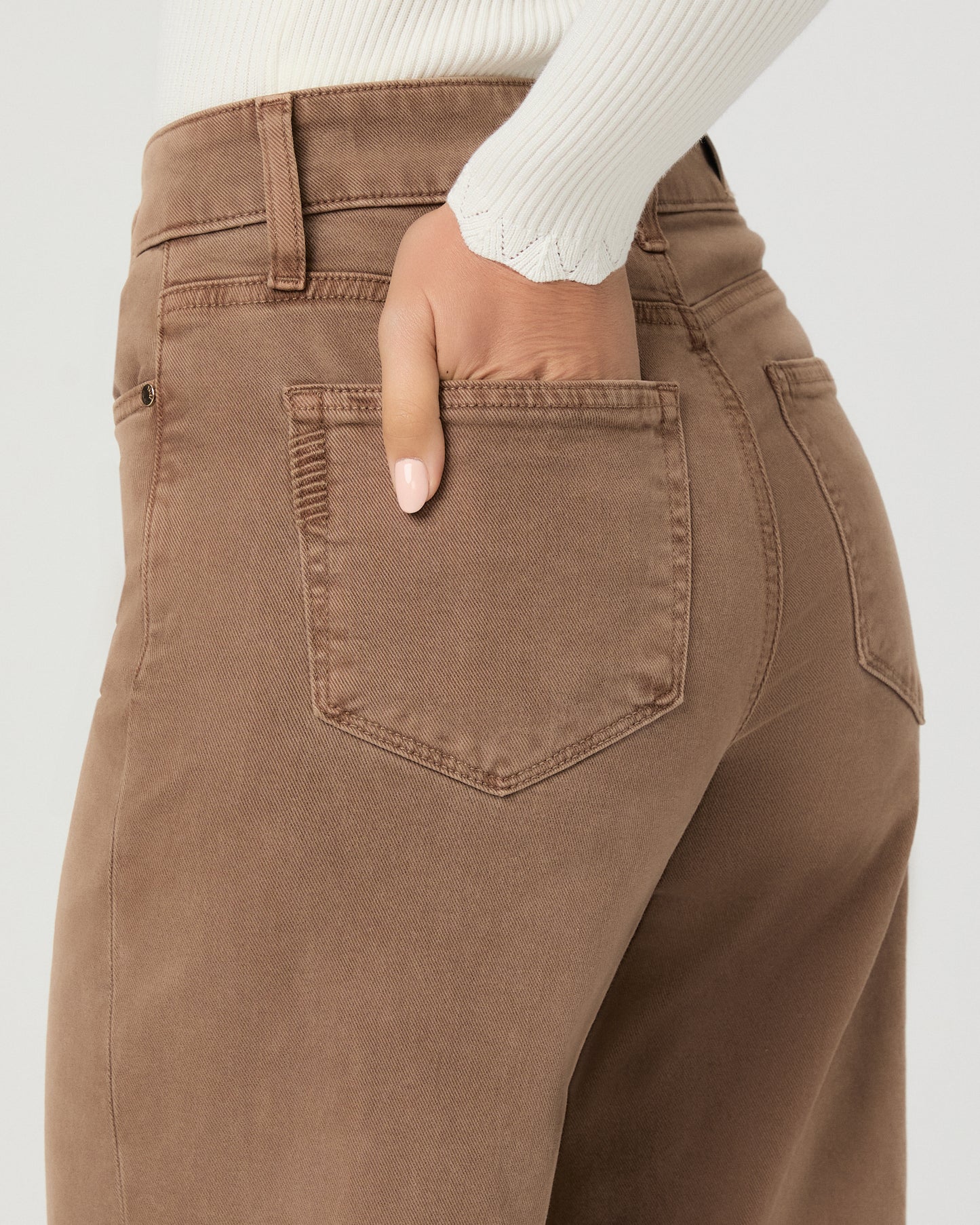 A woman modeling a pair of khaki pants with white long sleeve top and brown sandals against a light gray studio background.