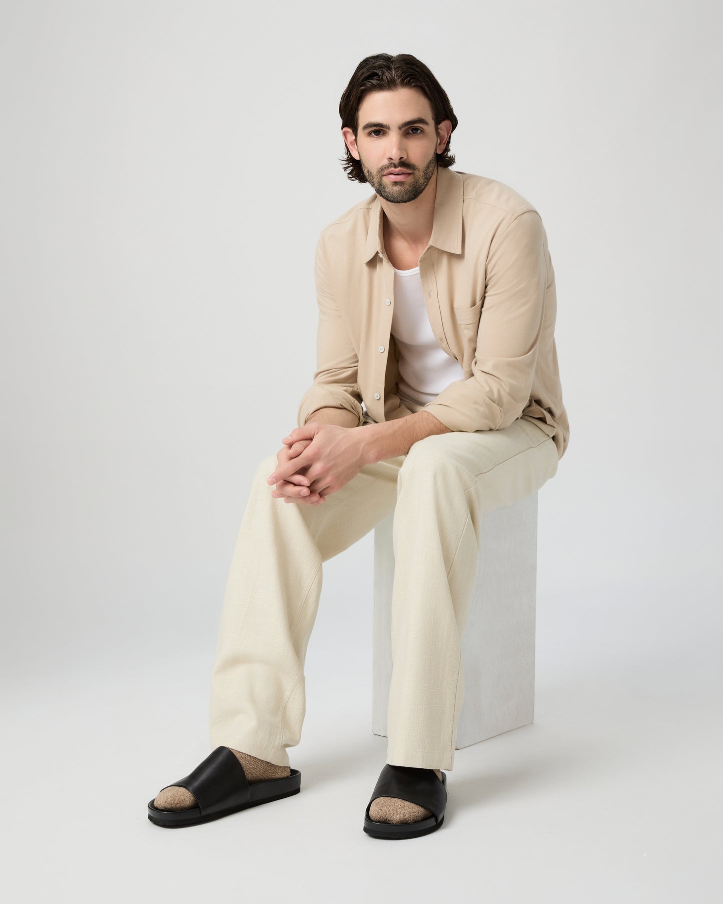 Man sitting on a white cube wearing a beige cardigan, white shirt, beige pants, and black sandals.