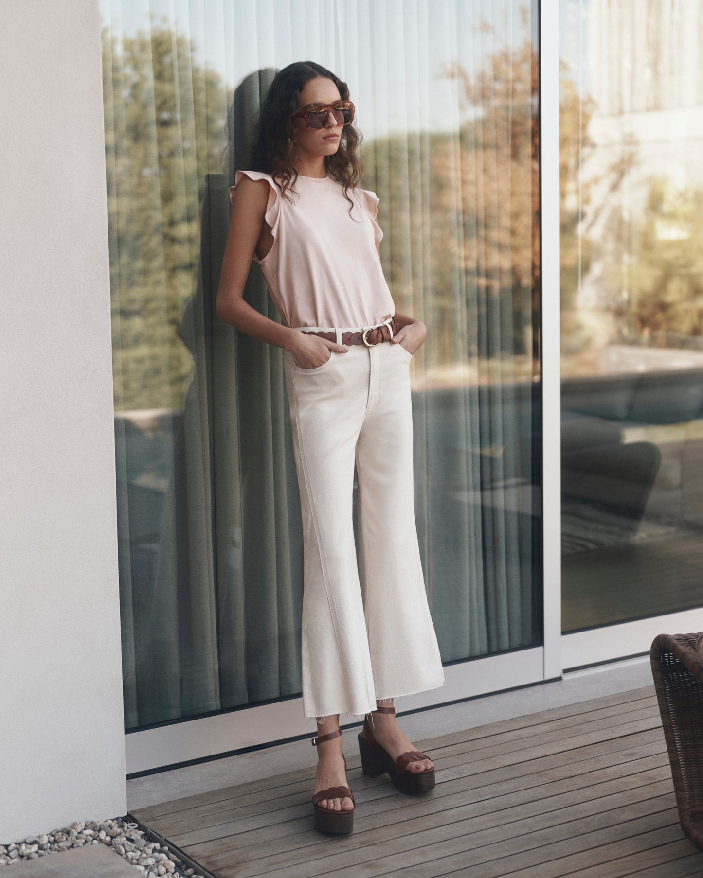 Woman in a pink top and white pants standing on a wooden deck with large glass doors in the background.