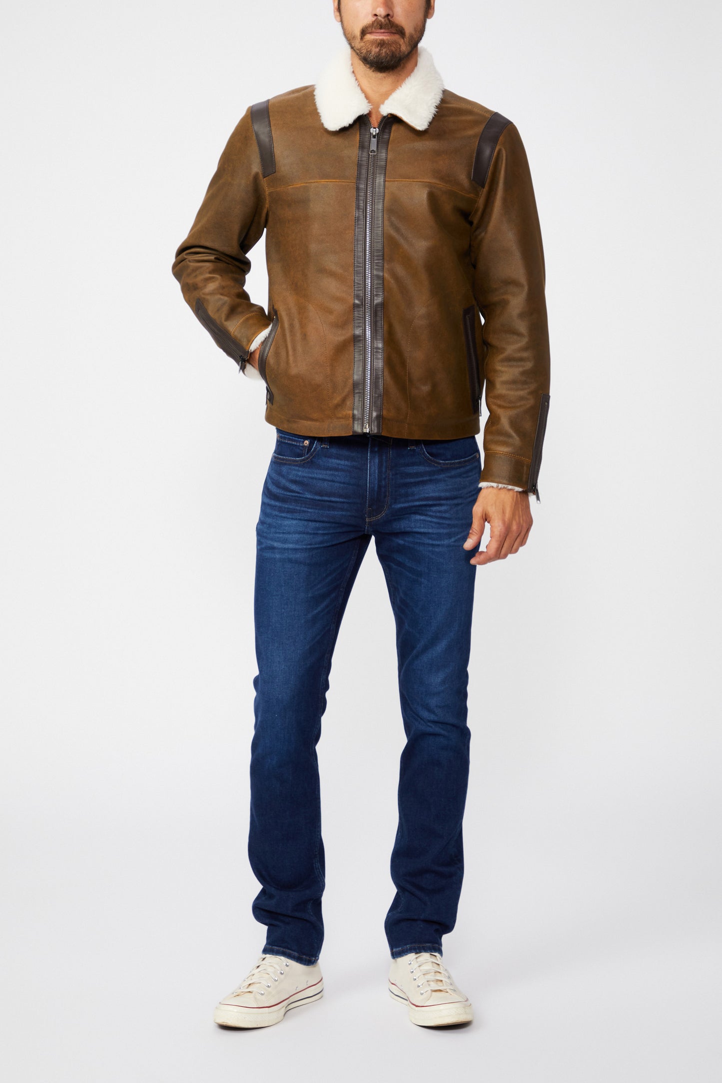 Man wearing a brown leather jacket with a white collar, blue jeans, and white sneakers on a white background