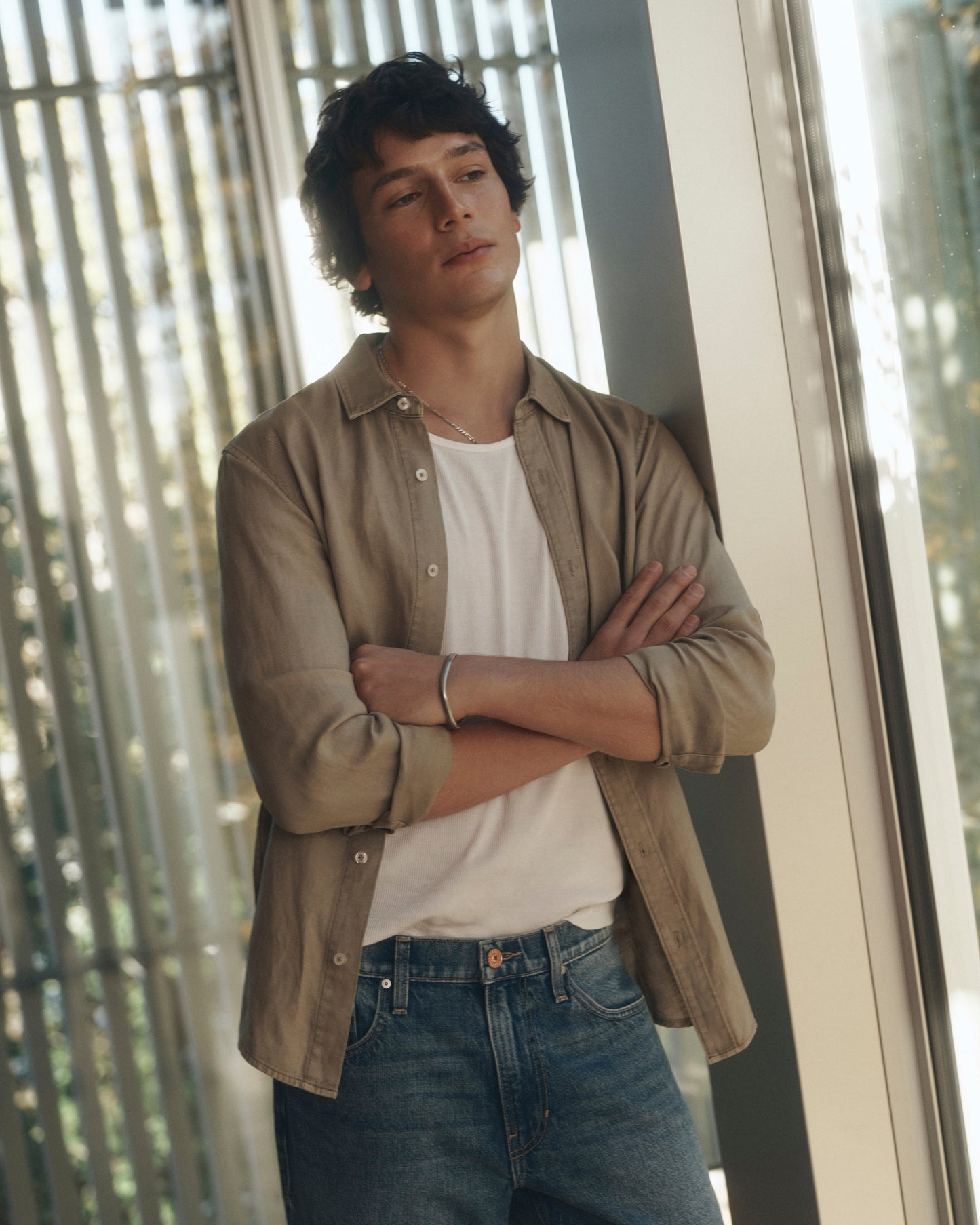 Man wearing a beige shirt over a white t-shirt and blue jeans, standing indoors with a window in the background.