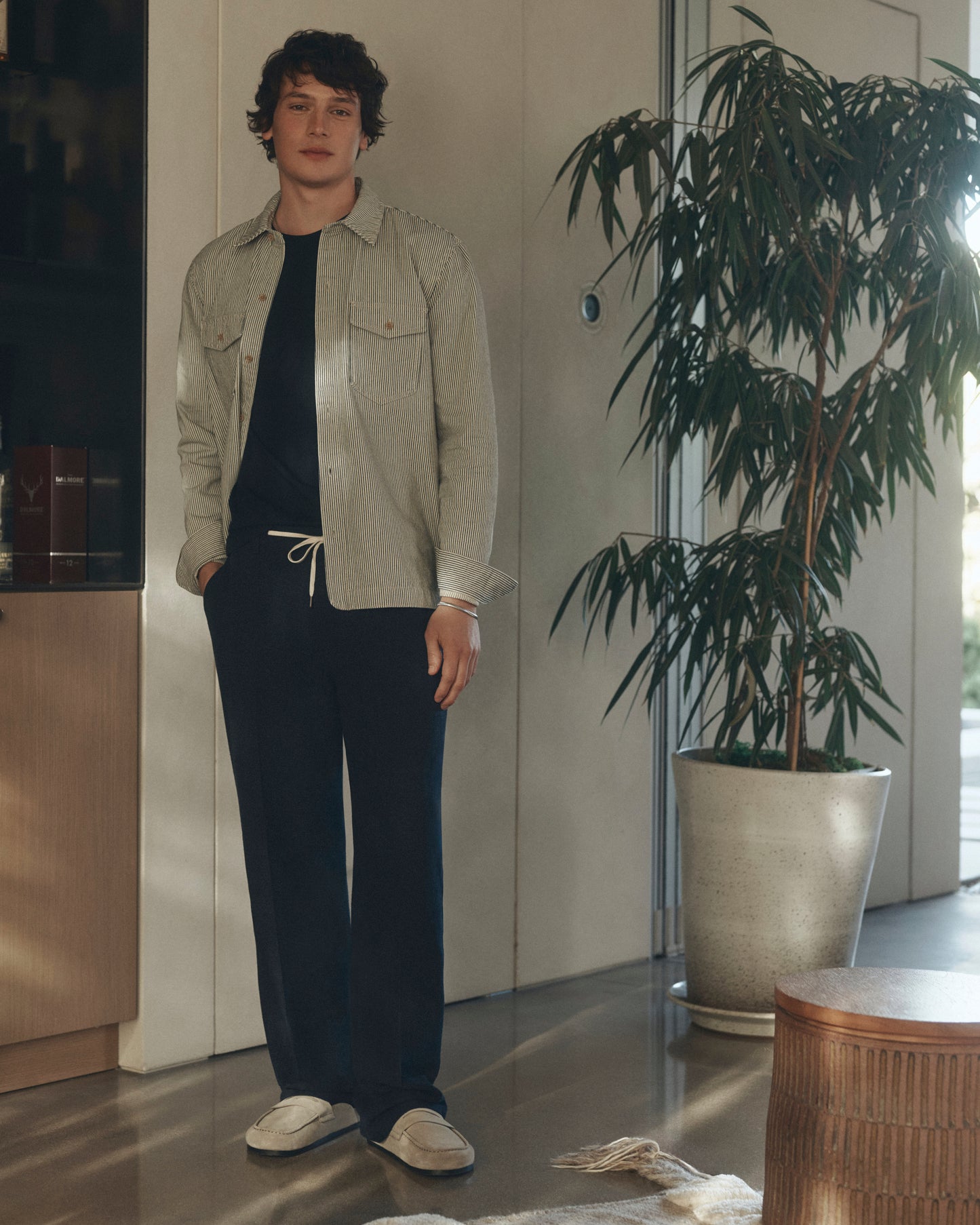 Person wearing a light jacket and dark pants standing indoors with a plant and furniture in the background