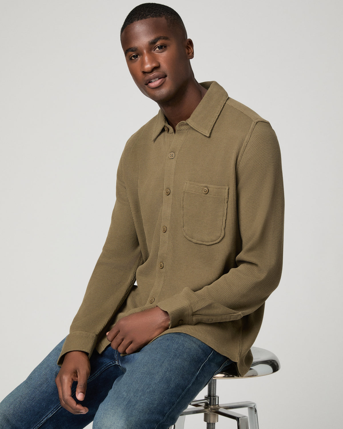 Close-up front view of a man in a deep olive green waffle-knit shirt, hand resting near the chest pocket, highlighting the textured fabric detail.