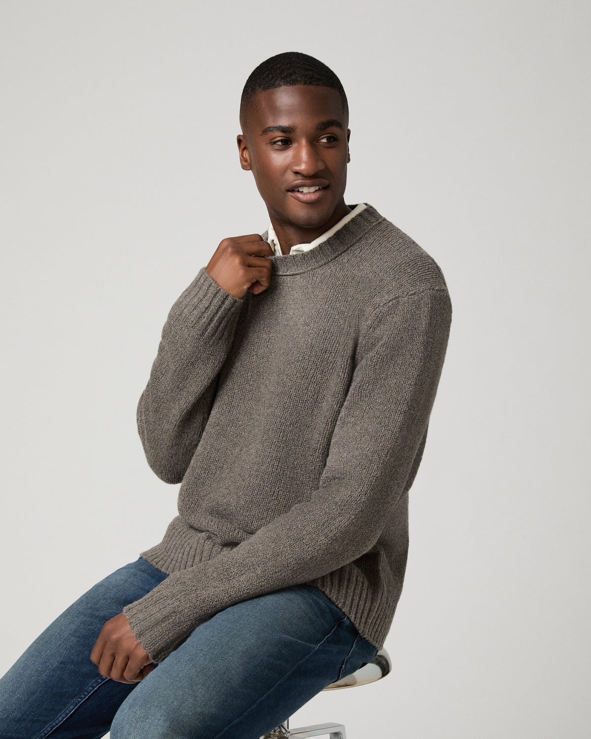 Seated three-quarter view of a man wearing the dark heather grey crewneck sweater over a white collared shirt, one hand at his collar, smiling and looking off to the side, paired with denim jeans.