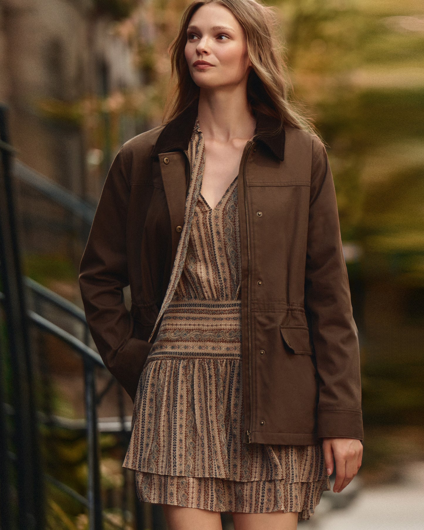 Woman wearing a brown jacket over a patterned dress in an outdoor setting
