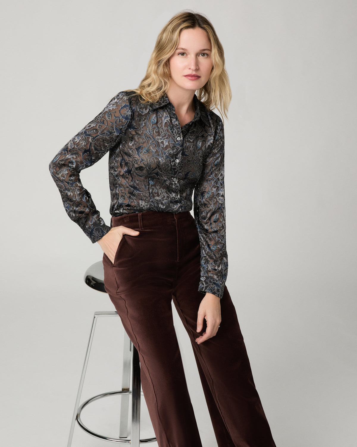 Women sitting down on a stool wearing a grey printed long sleeve top paired with suede burgundy jeans standing against a white background