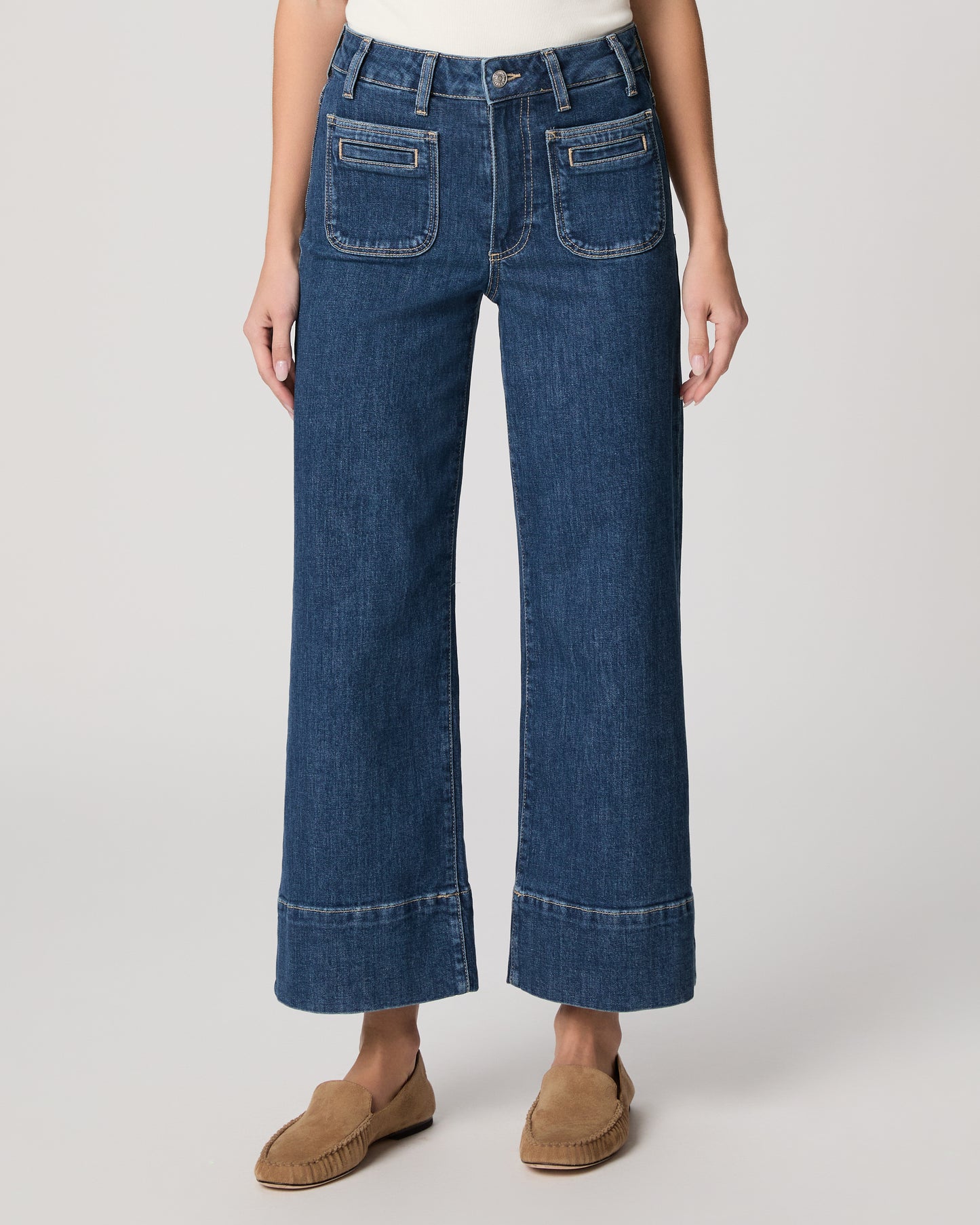 Woman wearing a white top and blue jeans paired with brown flats on a plain background