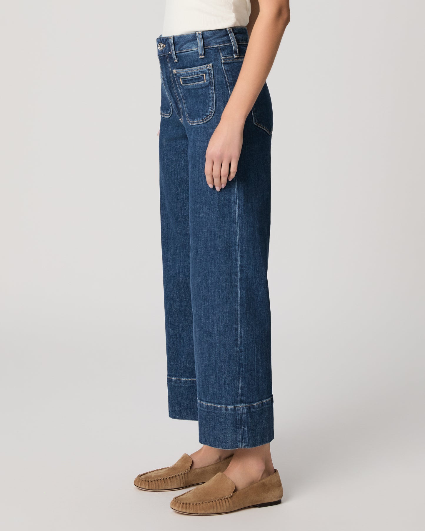 Woman wearing a white top and blue jeans paired with brown flats on a plain background