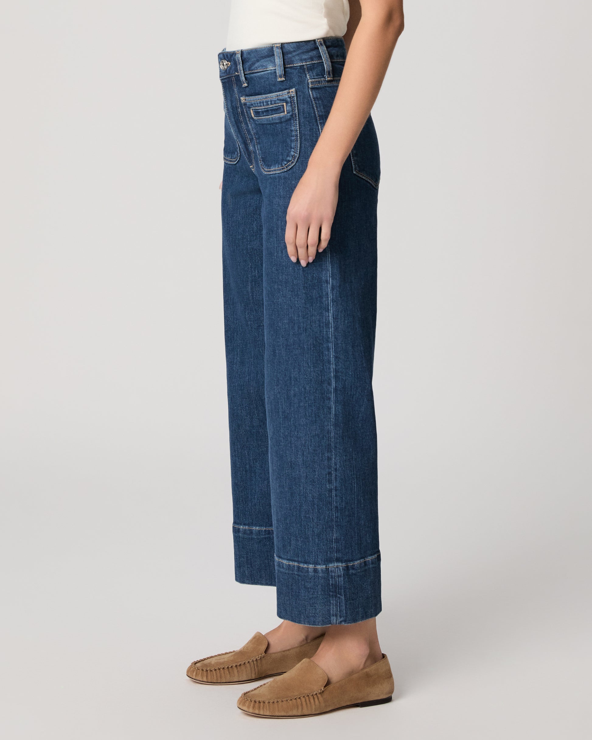 Woman wearing a white top and blue jeans paired with brown flats on a plain background