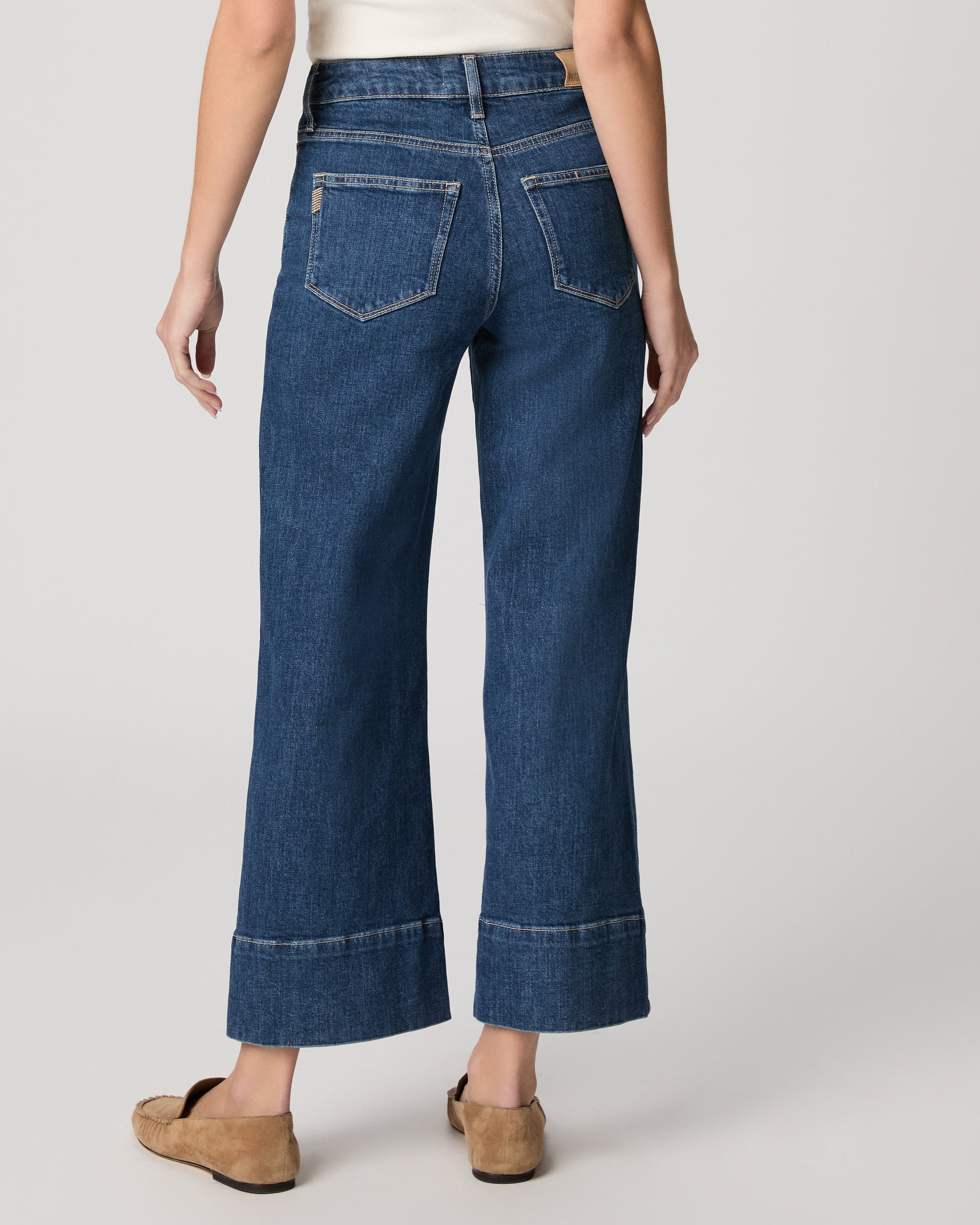 Woman wearing a white top and blue jeans paired with brown flats on a plain background