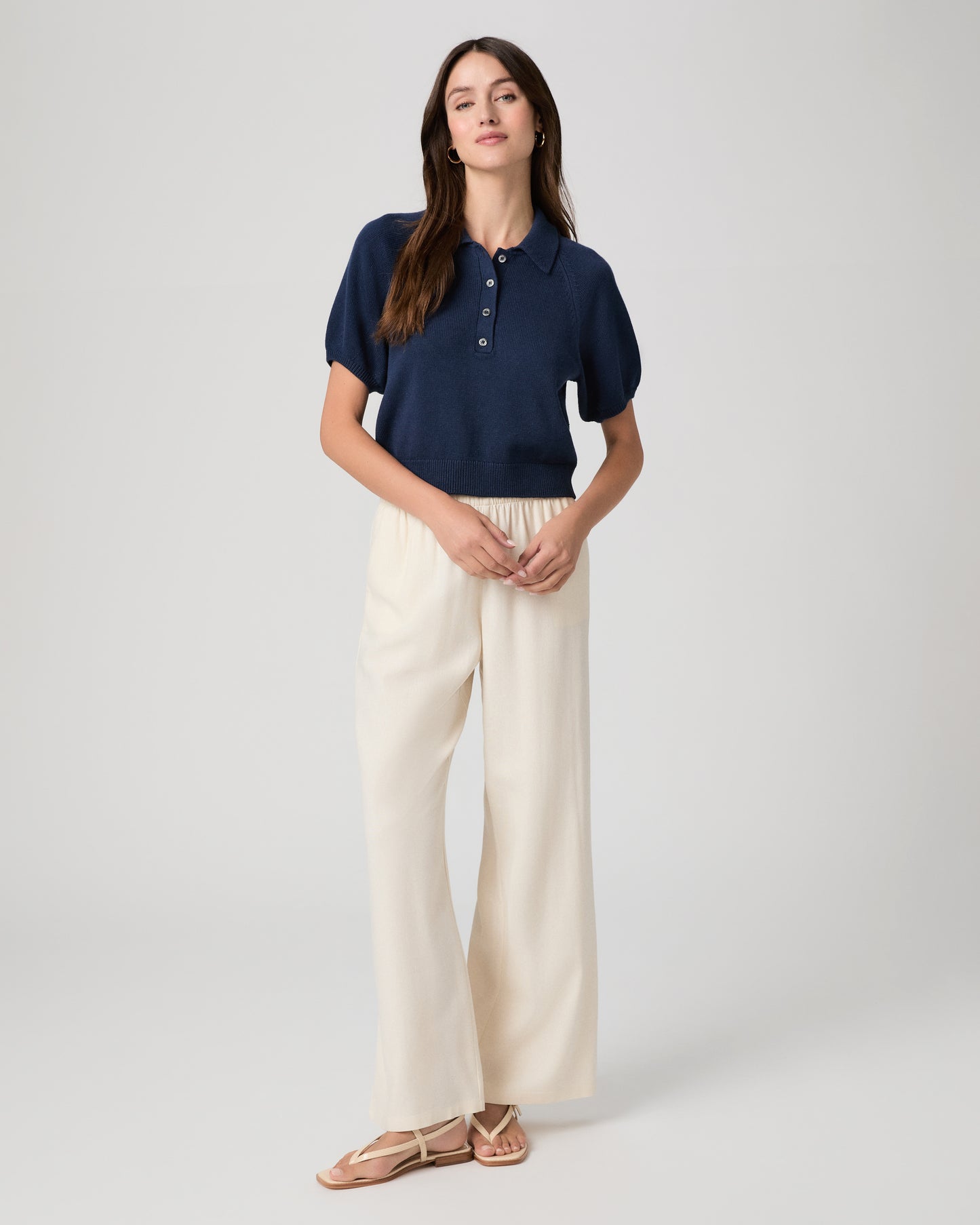 Woman wearing a navy blue polo shirt and white pants paired with white sandals on a light gray background