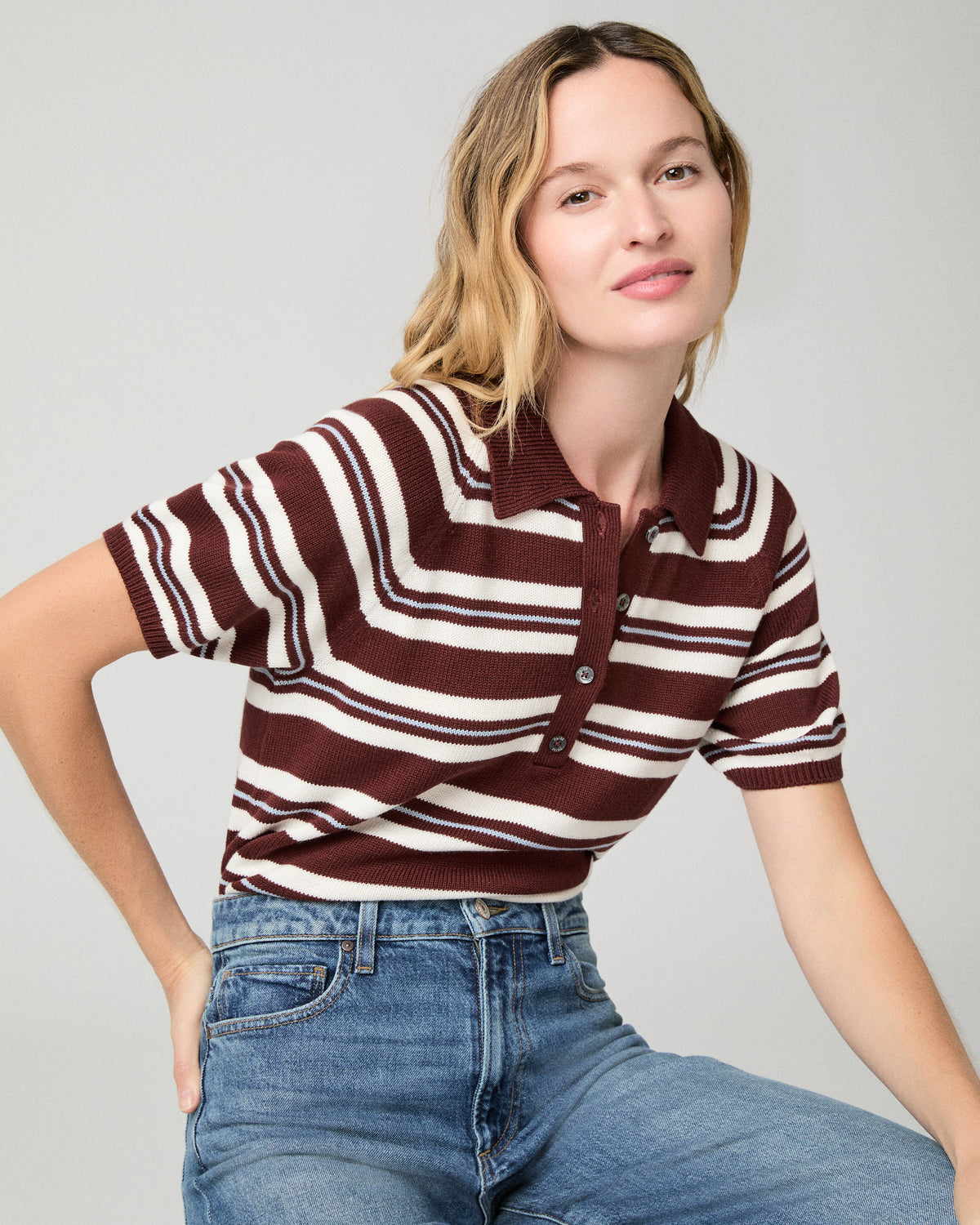 Women sitting on a stool wearing a white and brown stripe top and blue jeans against a white background