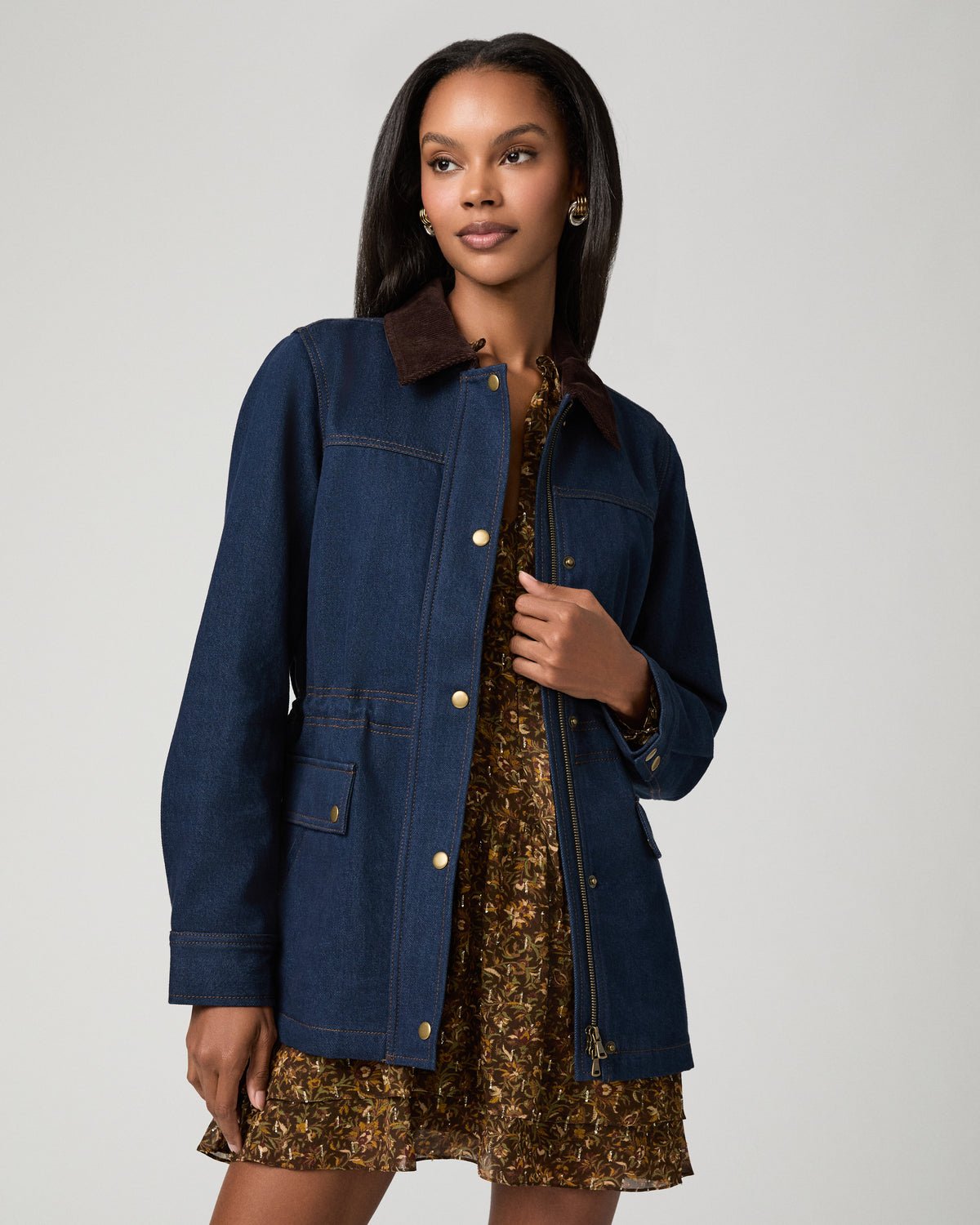 Full-length front view of a woman wearing a dark denim barn jacket with a brown corduroy collar and snap-front patch pockets, worn open over a brown floral-print dress.