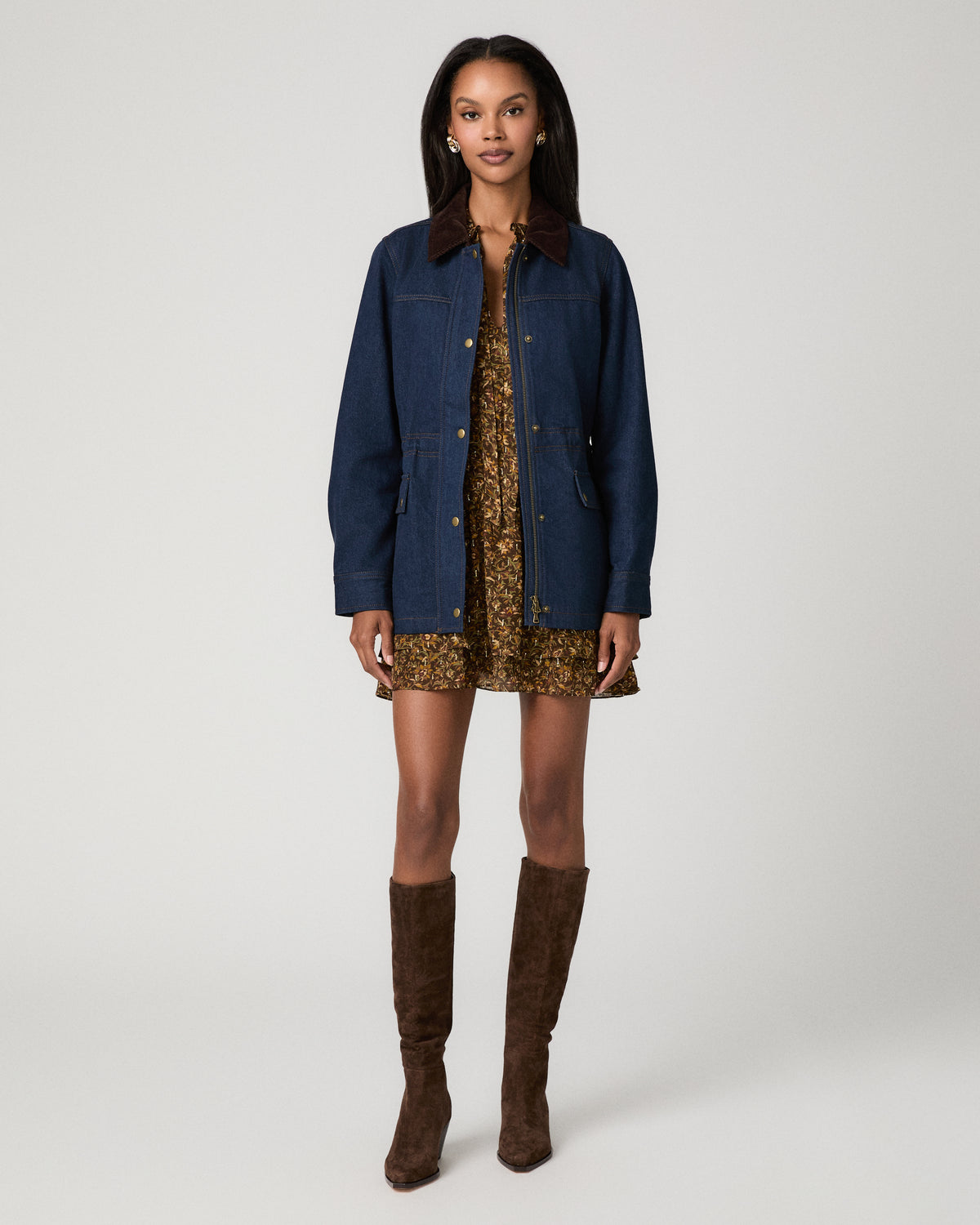 Full-length front view of a woman wearing a dark denim barn jacket snapped closed over a brown floral mini dress, paired with tall brown suede boots.