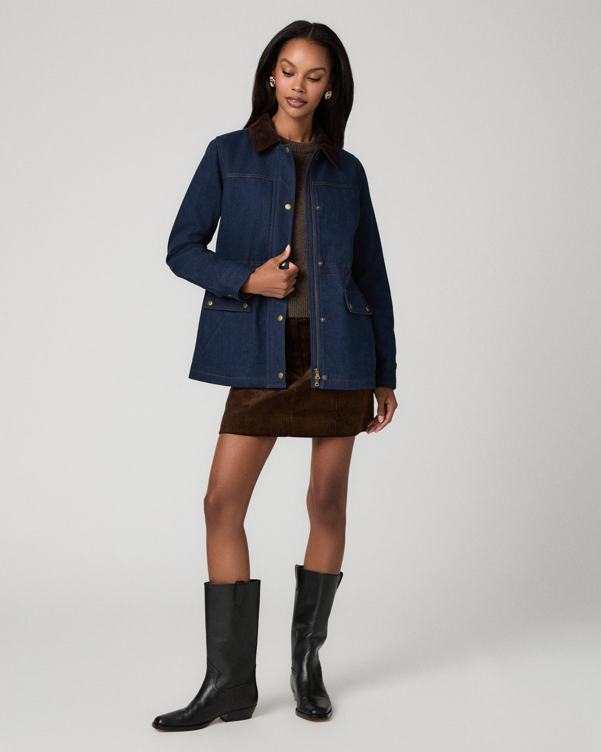 Full-length front view of a woman wearing a dark denim barn jacket open over a brown corduroy mini skirt and top, with black knee-high boots.