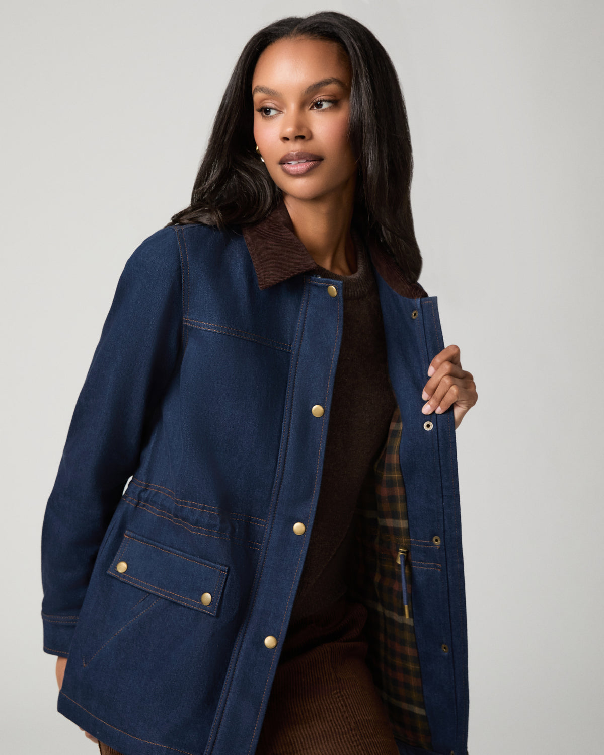 Close-up front view of a woman in a dark denim barn jacket with a brown corduroy collar and snap-front placket, one hand tucked near the collar.
