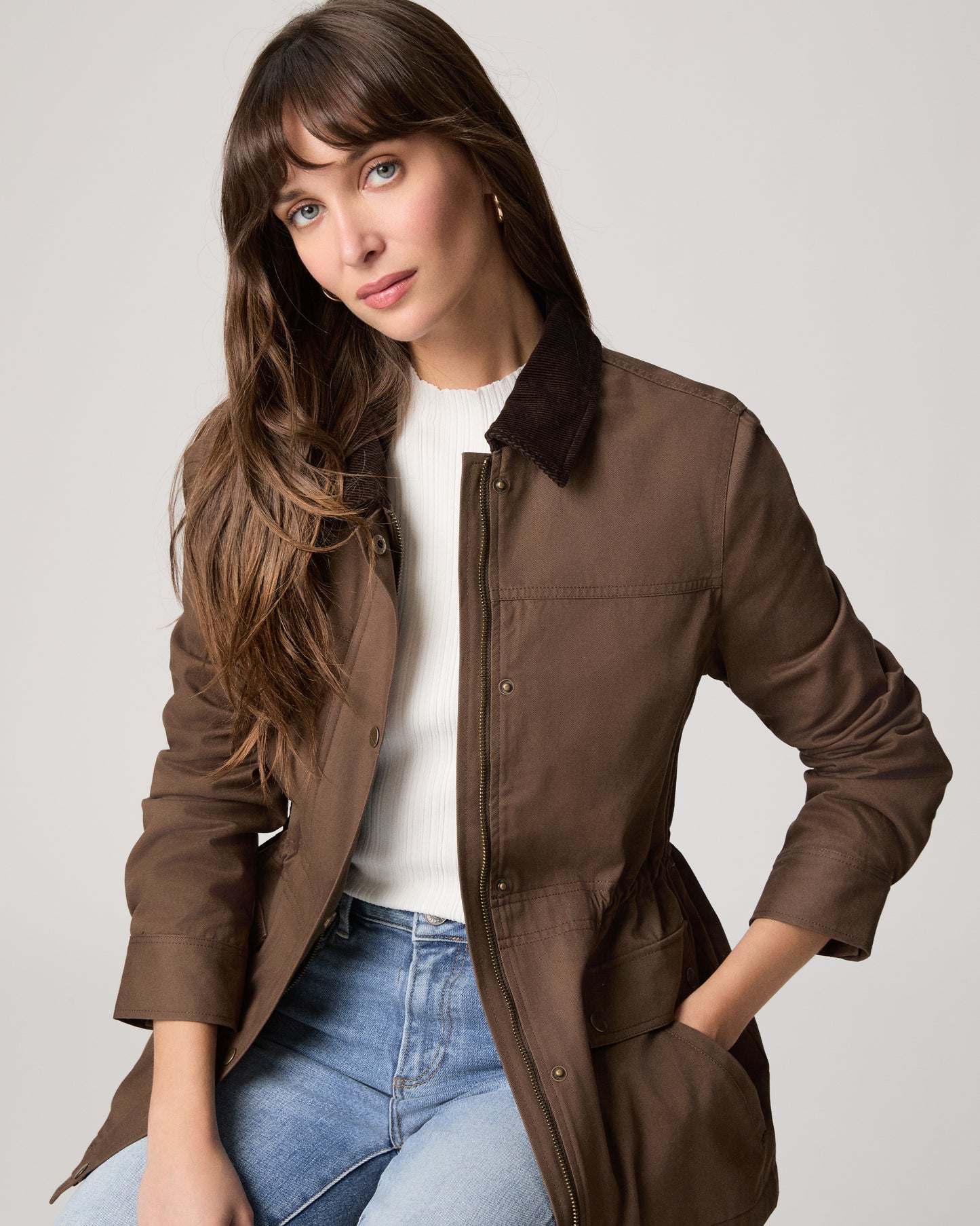 Woman wearing a brown jacket over a white shirt and blue jeans on a light gray background