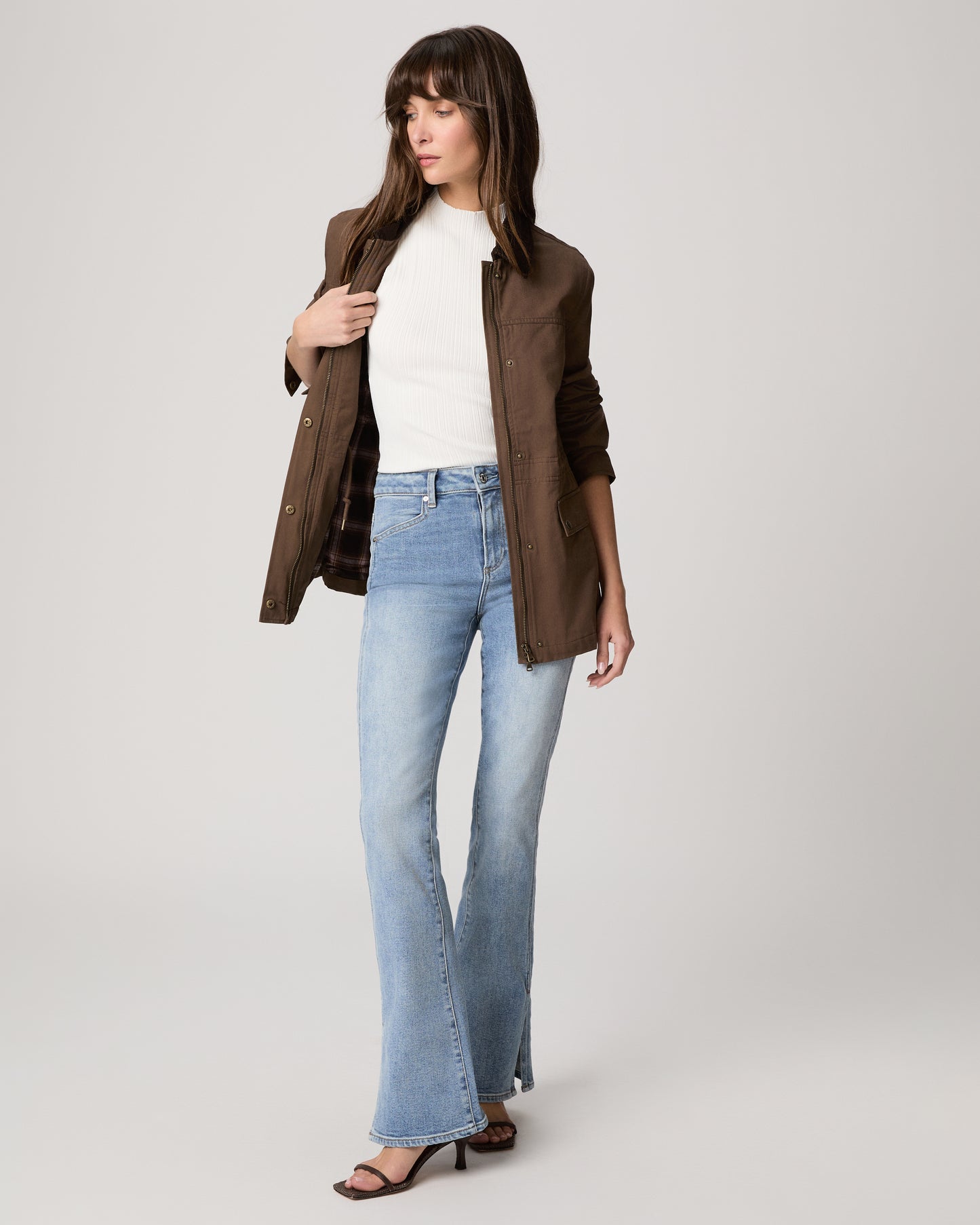 Woman wearing a brown jacket, white shirt, and blue jeans on a light gray background