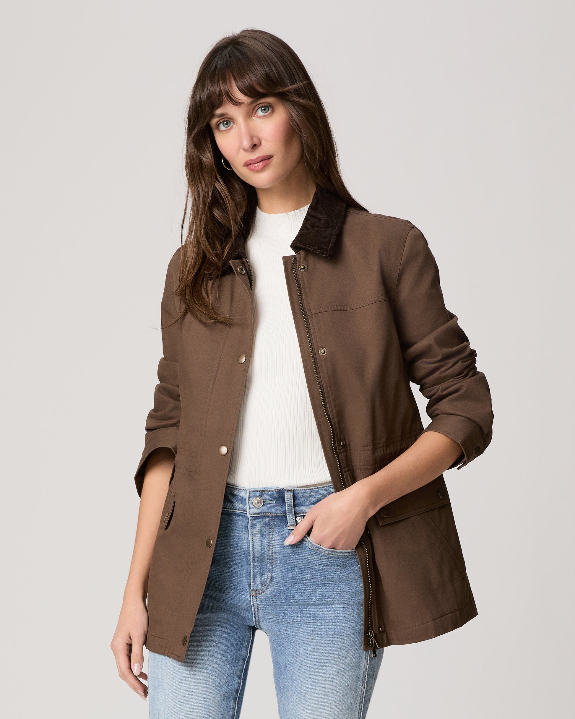 Woman wearing a brown jacket with a white shirt and blue jeans on a light gray background