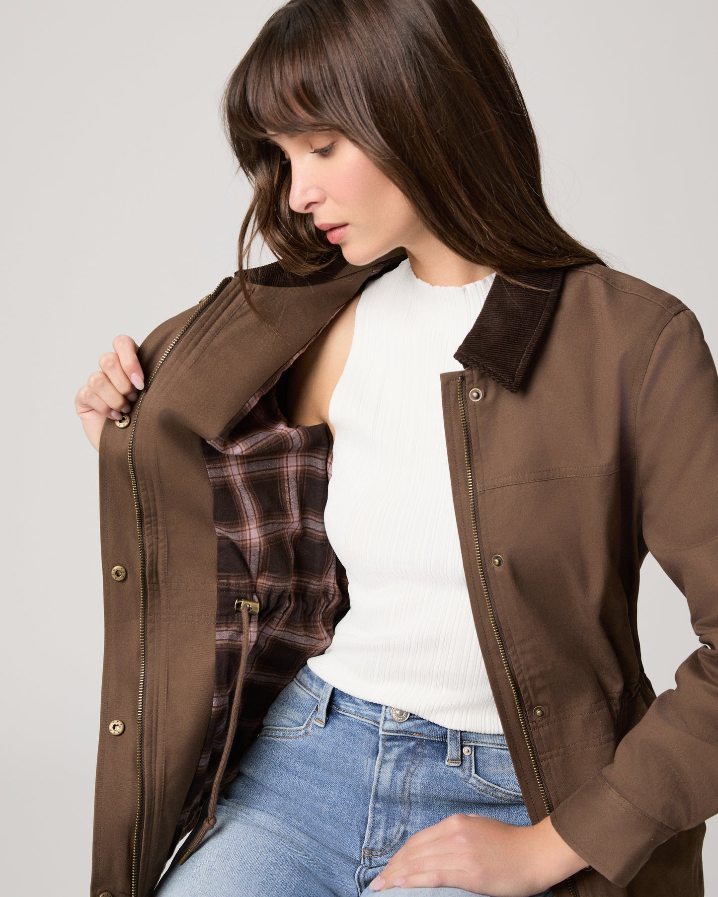 Woman wearing a brown jacket over a plaid shirt with a plain background